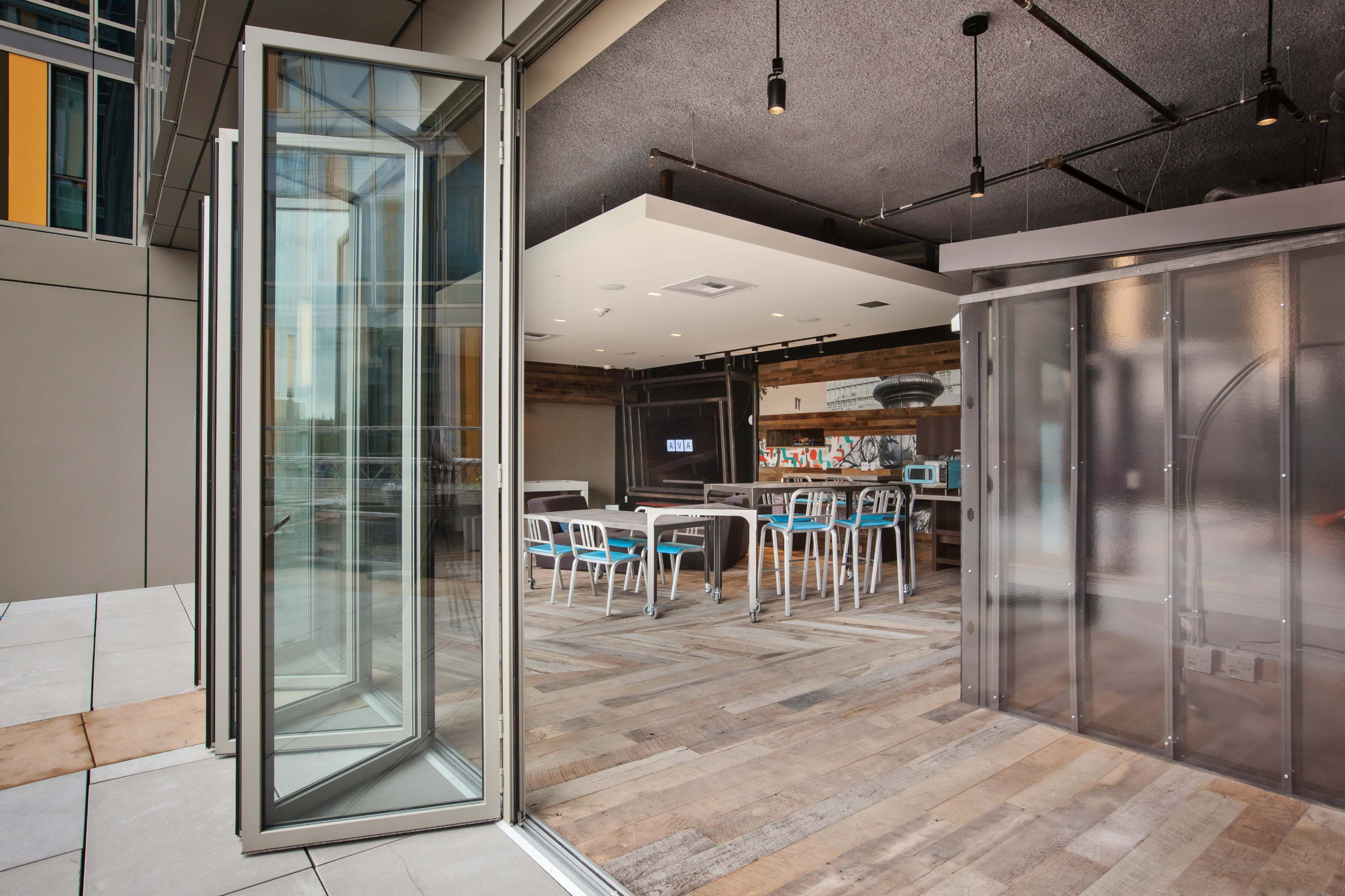 A modern indoor meeting space with large glass doors open to an outdoor area, featuring a wooden floor and a mix of tables and chairs.