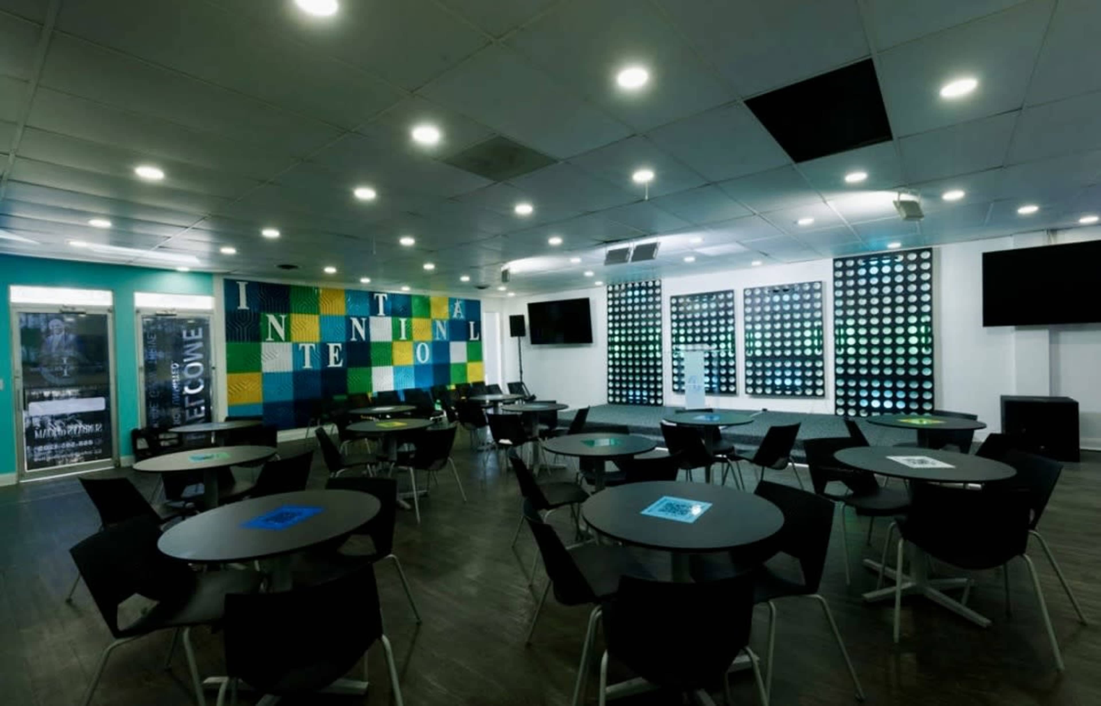The image shows a modern meeting room with several round tables and a colorful wall featuring large letters.