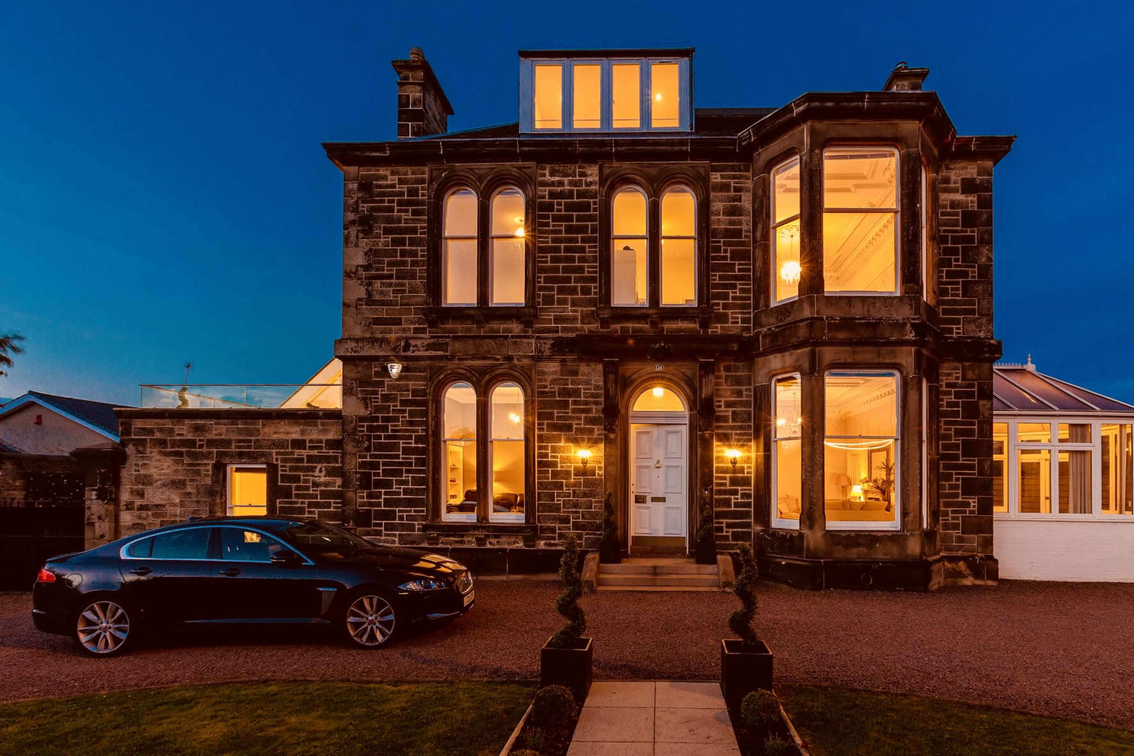 A large, historic stone house with multiple windows and a dark-colored car parked in front.