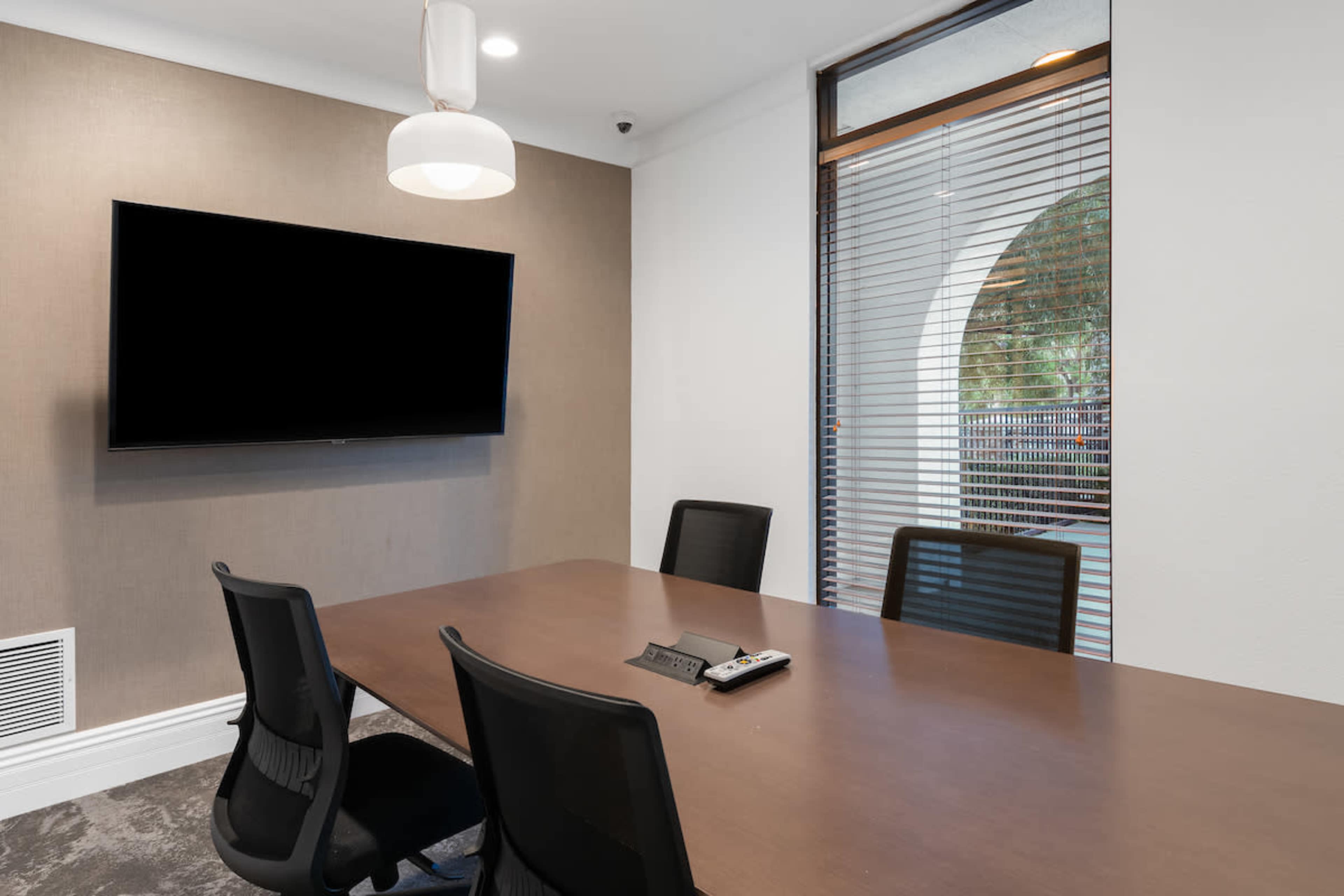 A conference room features a large table with four chairs, a wall-mounted television, and a window with blinds.