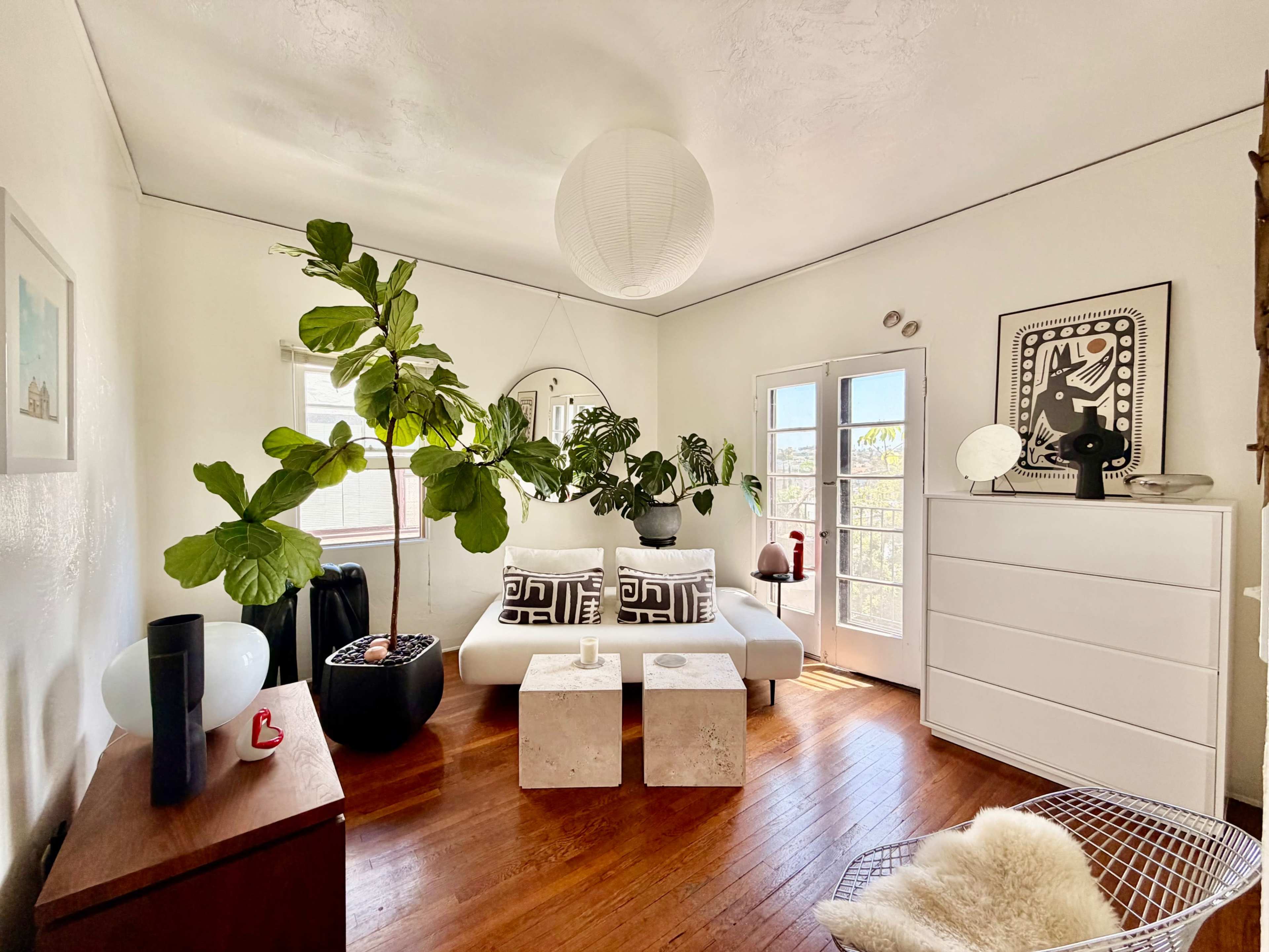 The image shows a bright living room featuring a white sofa, a pair of concrete side tables, and large indoor plants, with French doors leading outside.