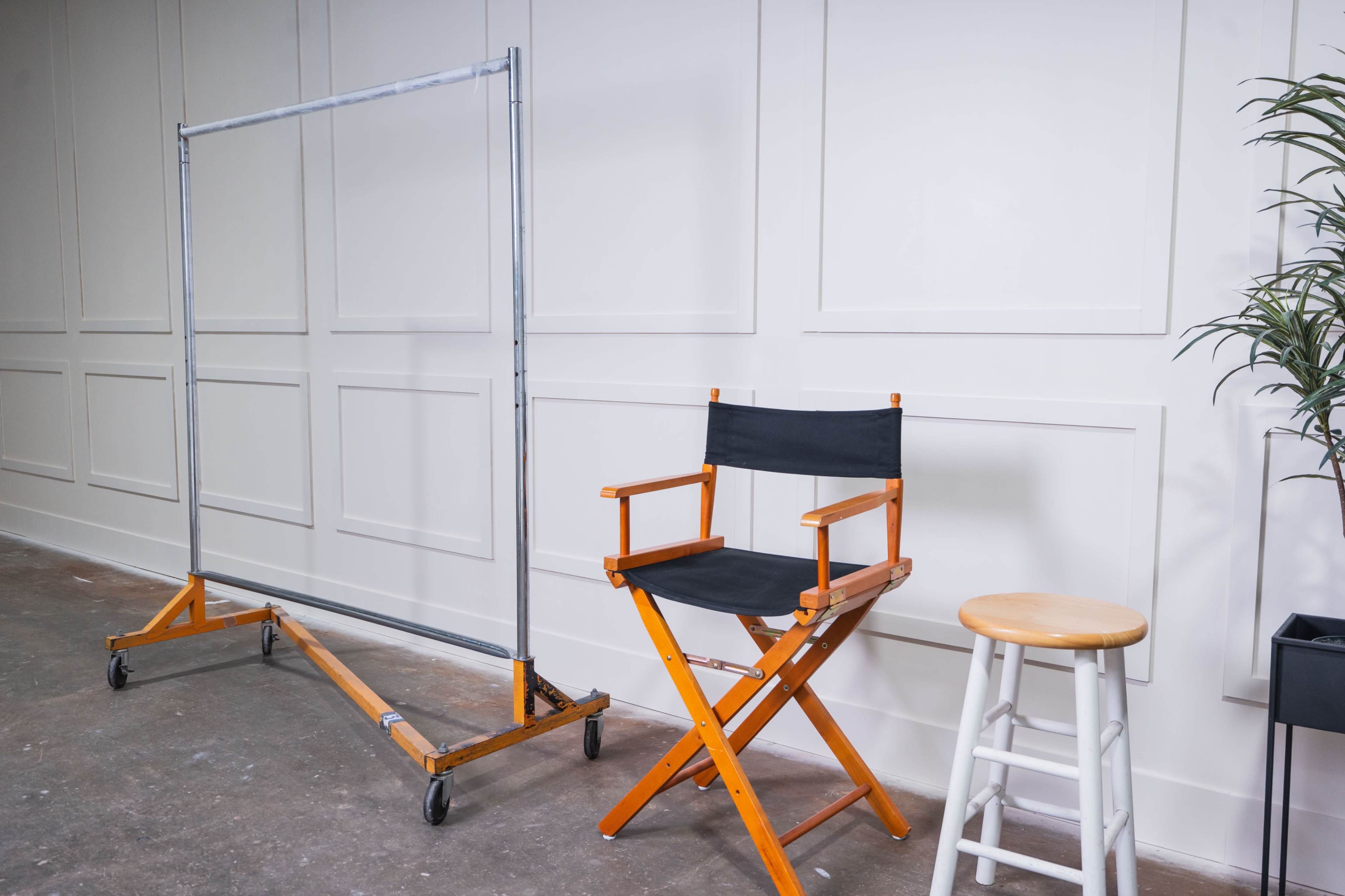 The image shows a wooden director's chair next to a white stool and a metal clothing rack in a minimalist interior space.
