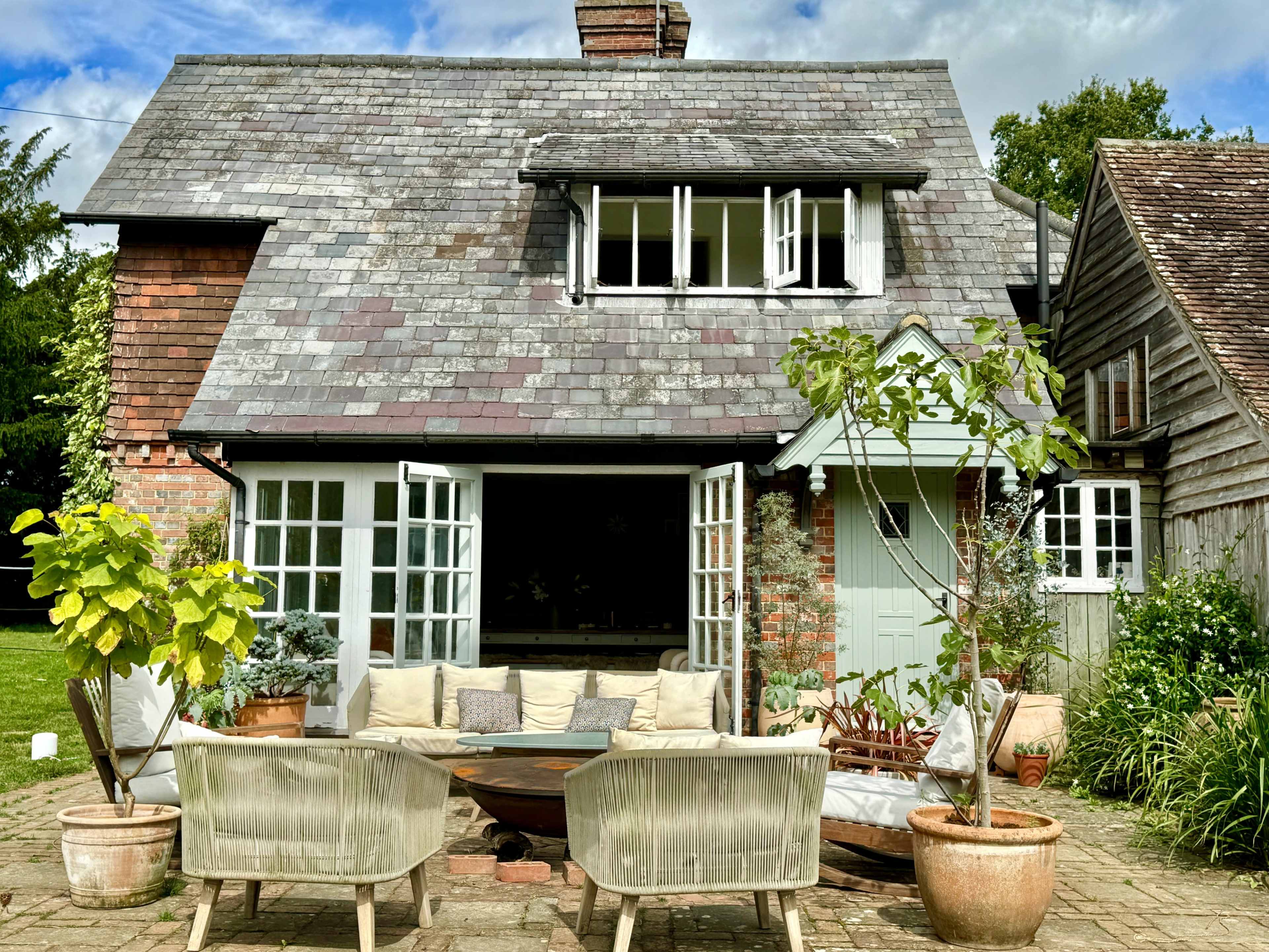 A rustic house with a slate roof and an outdoor seating area featuring two armchairs and a central table, surrounded by green plants.