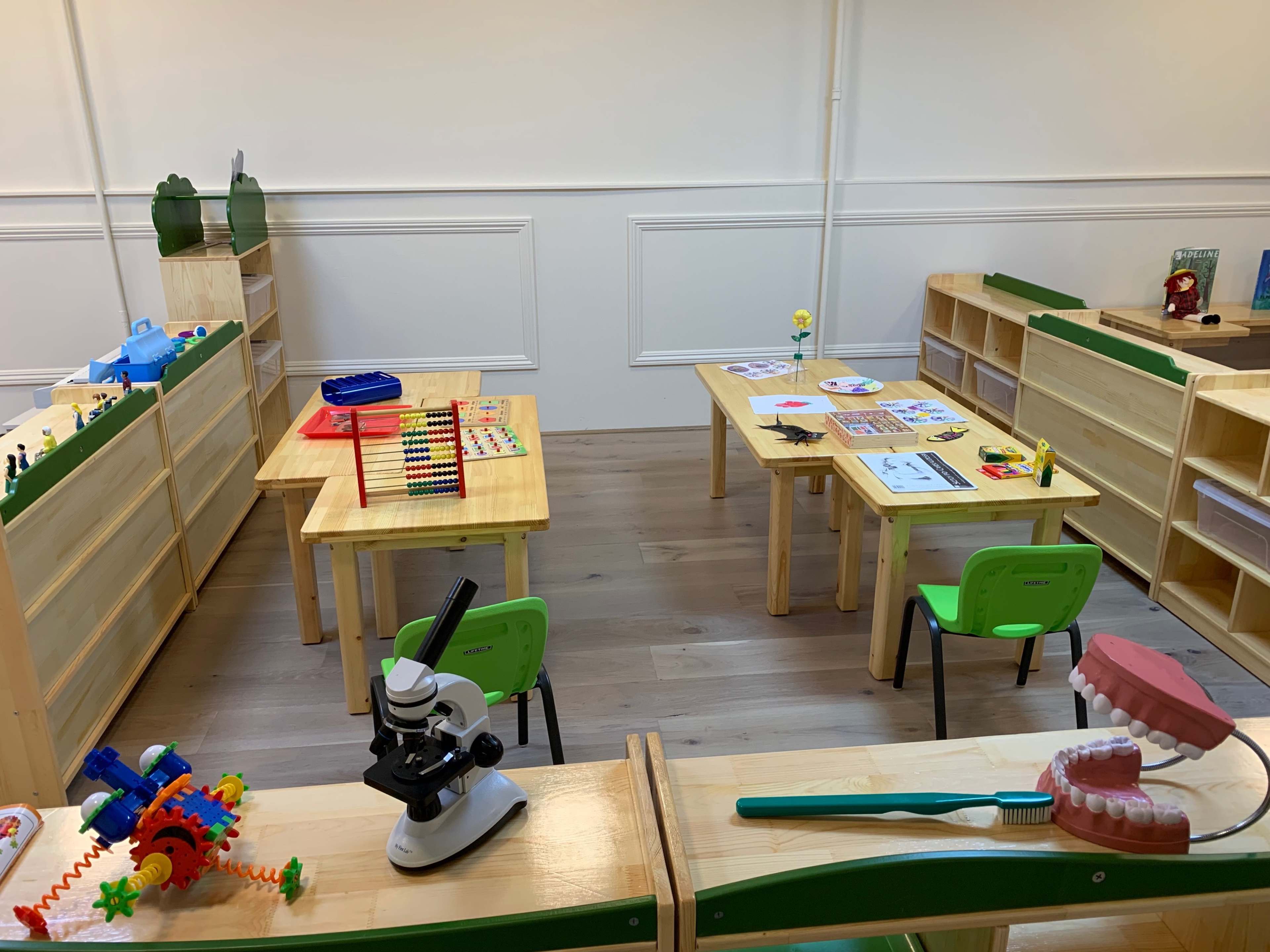 A bright classroom with wooden tables and shelves, featuring educational materials like a microscope, toys, and posters.