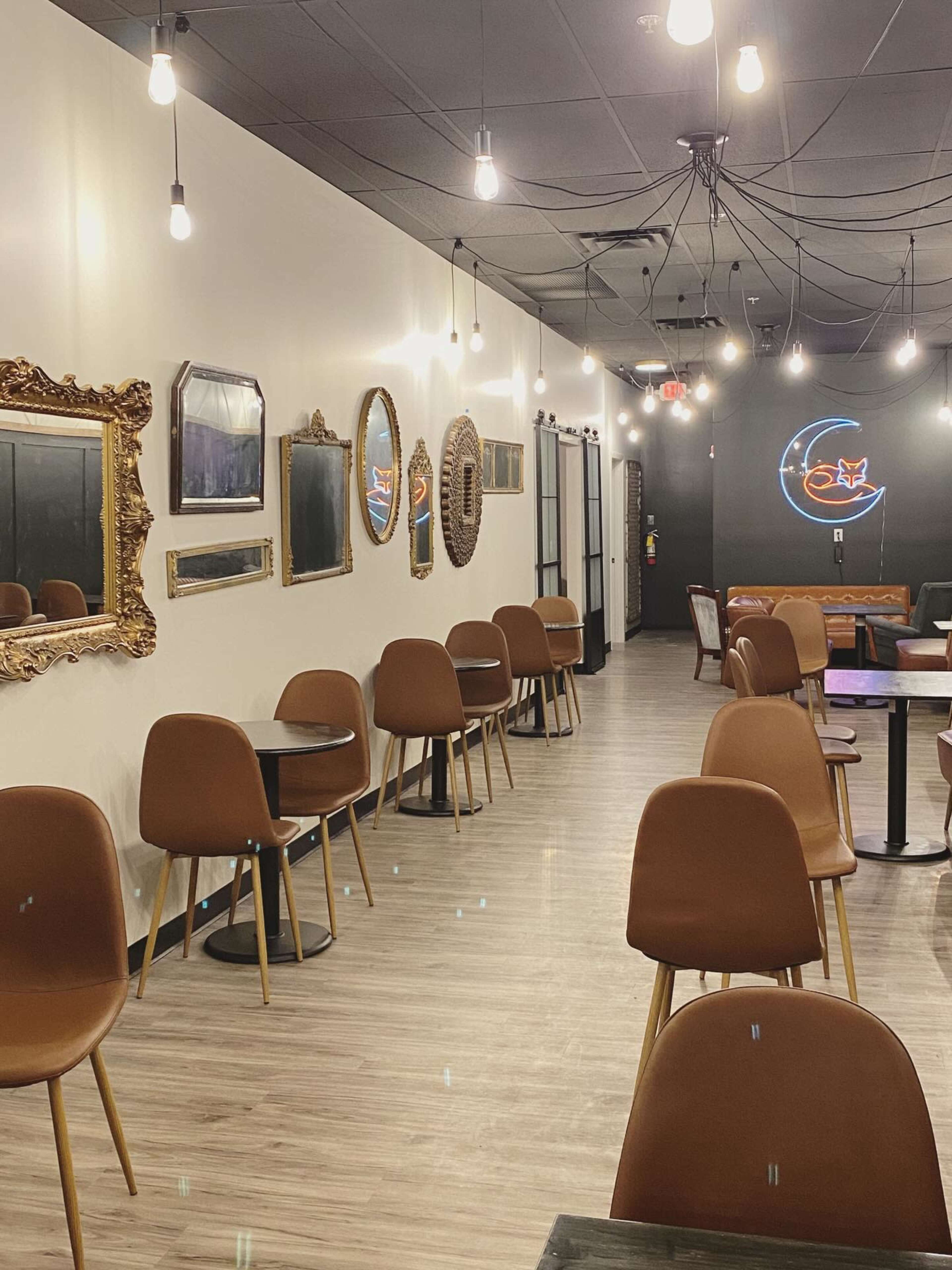 The image shows a modern café interior featuring brown chairs and tables, illuminated by pendant lights, with framed mirrors and artwork on the walls.