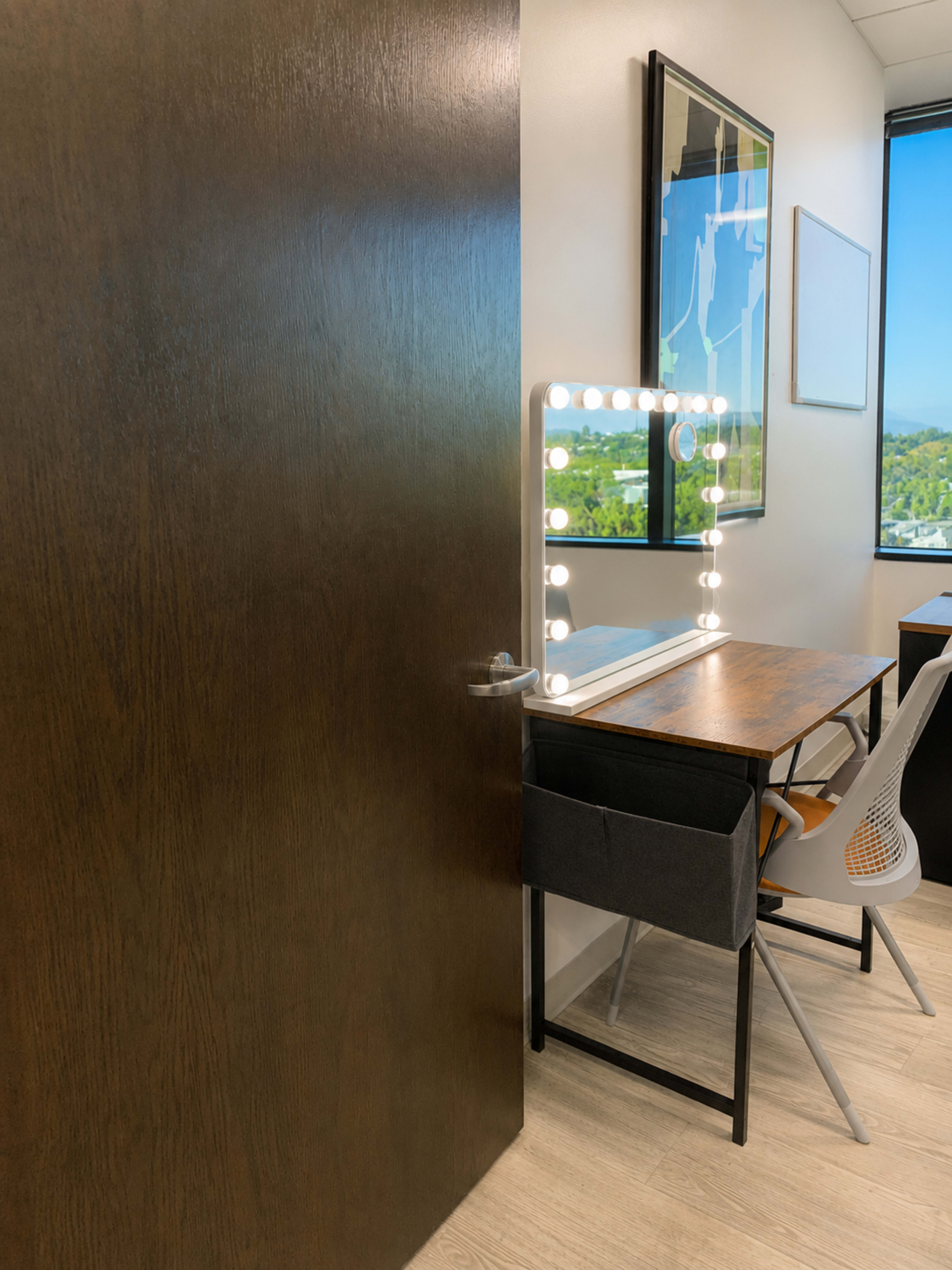 The image shows a modern workspace featuring a desk with a large illuminated mirror and a chair, viewed from an open door.