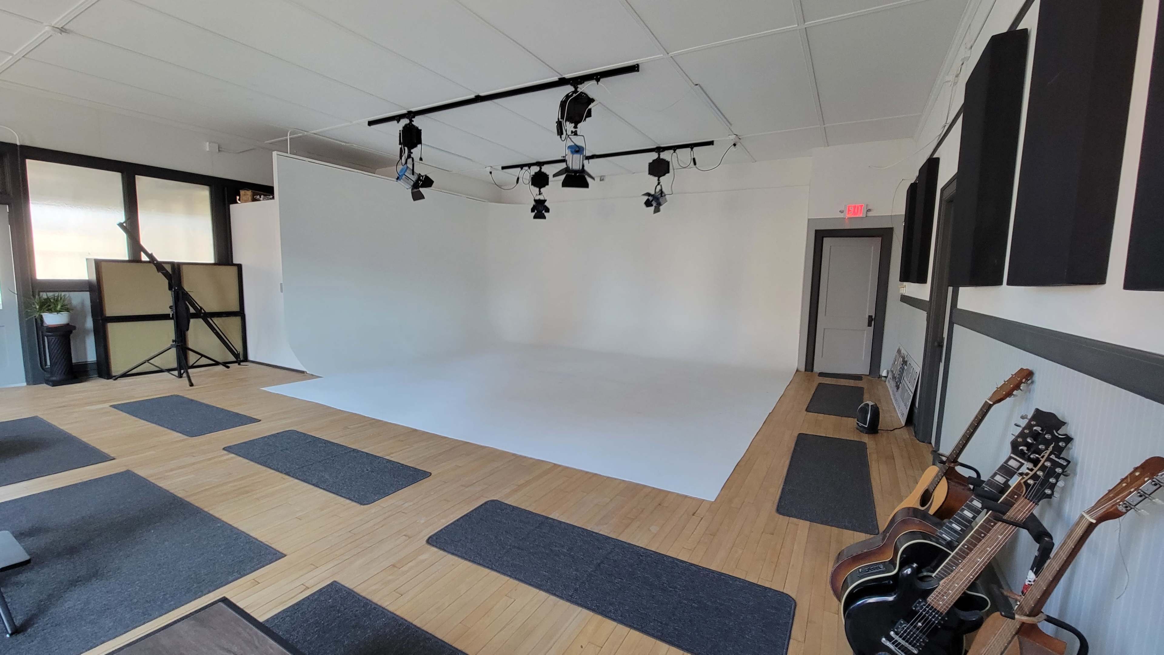 A spacious photography studio with a white backdrop, track lighting, and wooden flooring, featuring guitars in the corner.