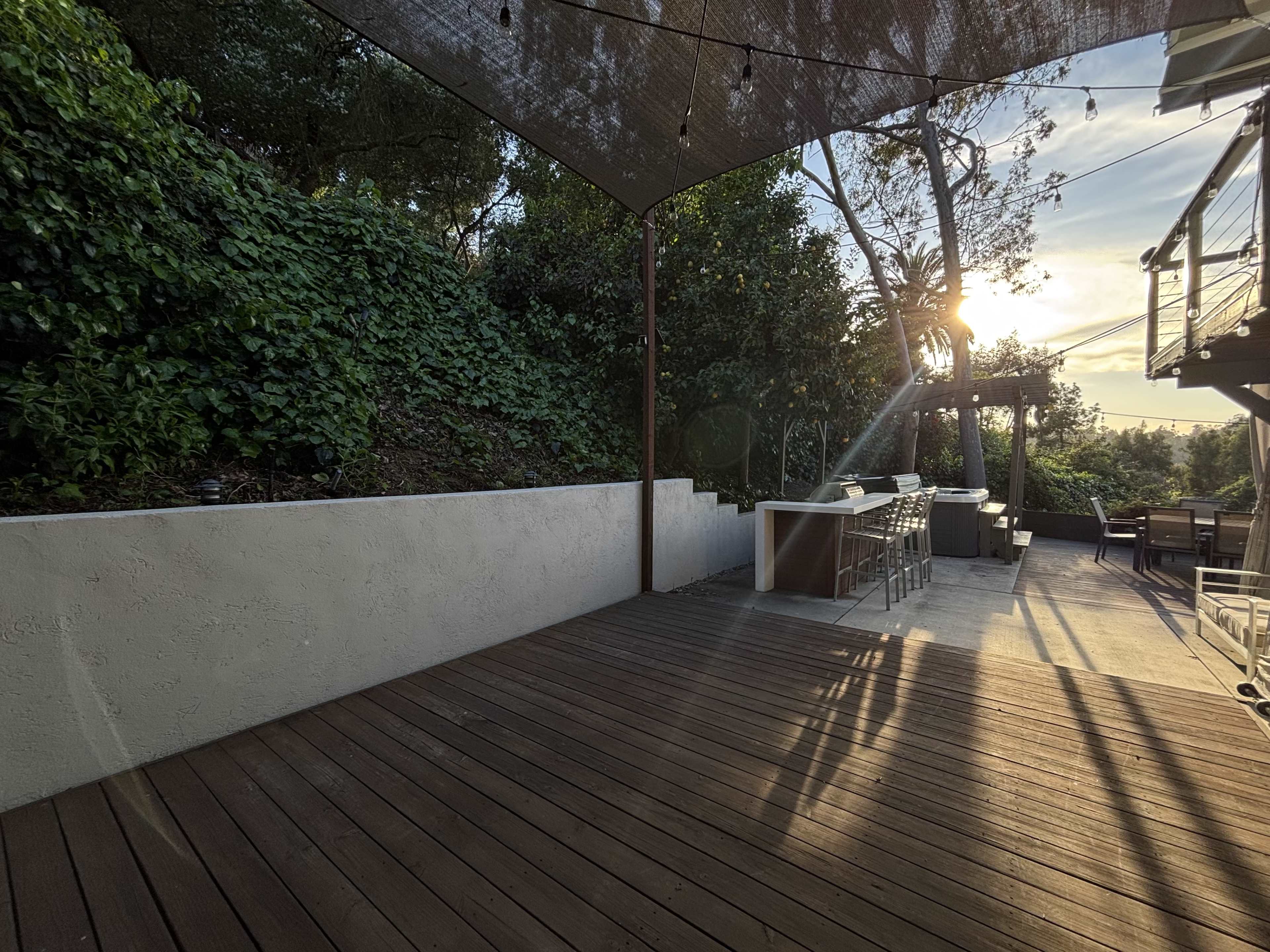 The image shows a wooden deck area featuring an outdoor kitchen and seating arrangement, surrounded by greenery and illuminated by sunset.