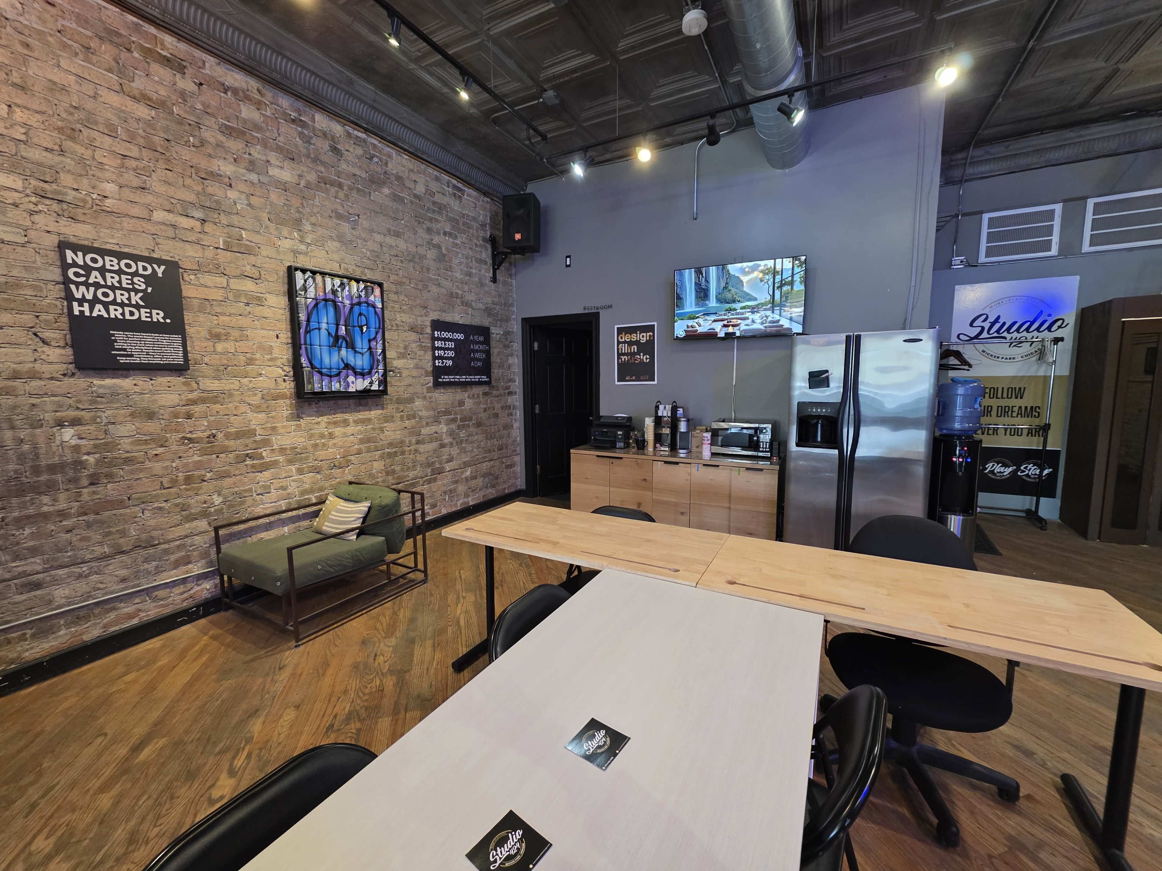 The image shows a contemporary office or co-working space with brick walls, a kitchenette area, and tables arranged for collaborative work.