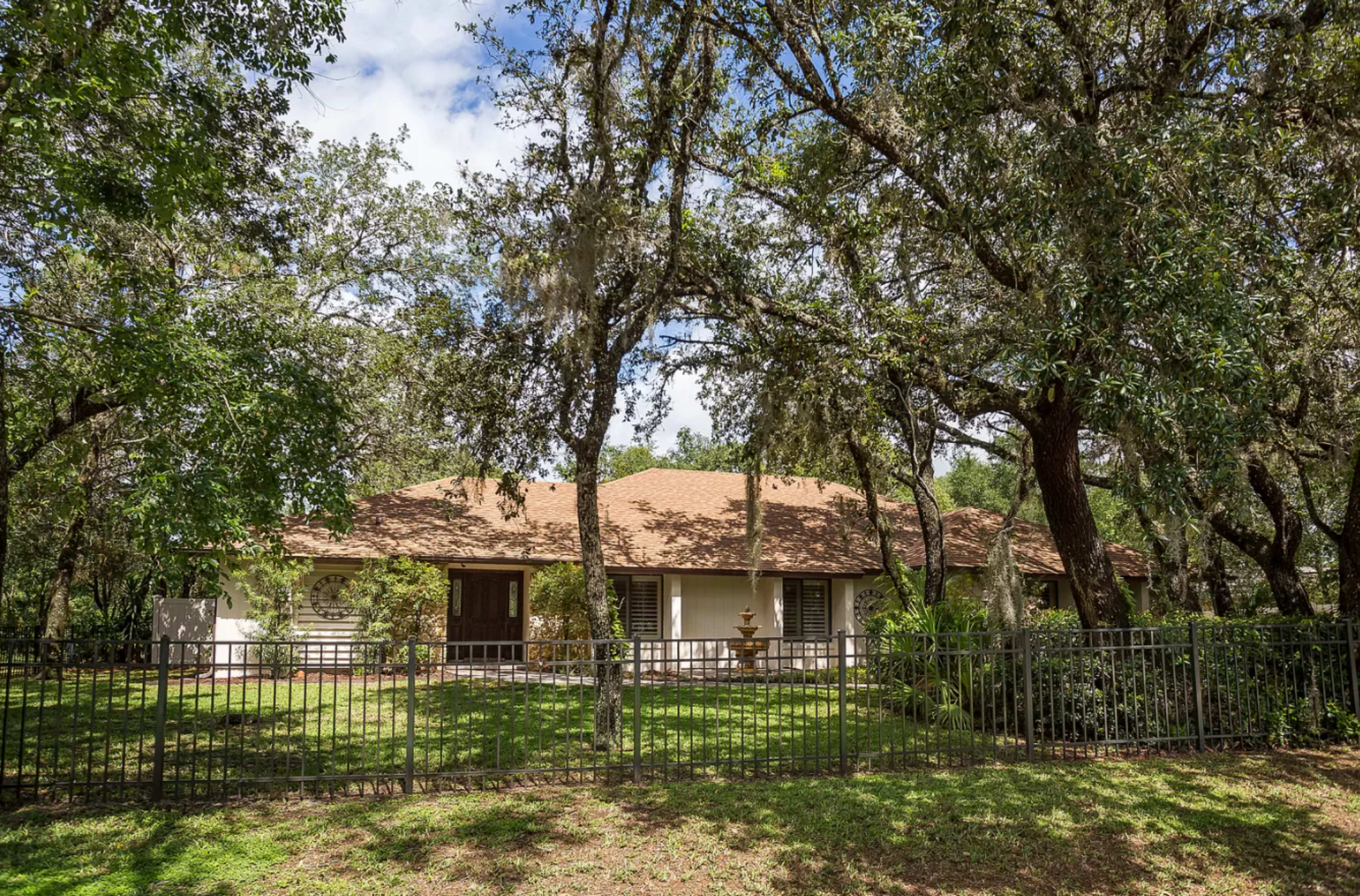 A single-story house with a brown roof is surrounded by trees and a black wrought iron fence.