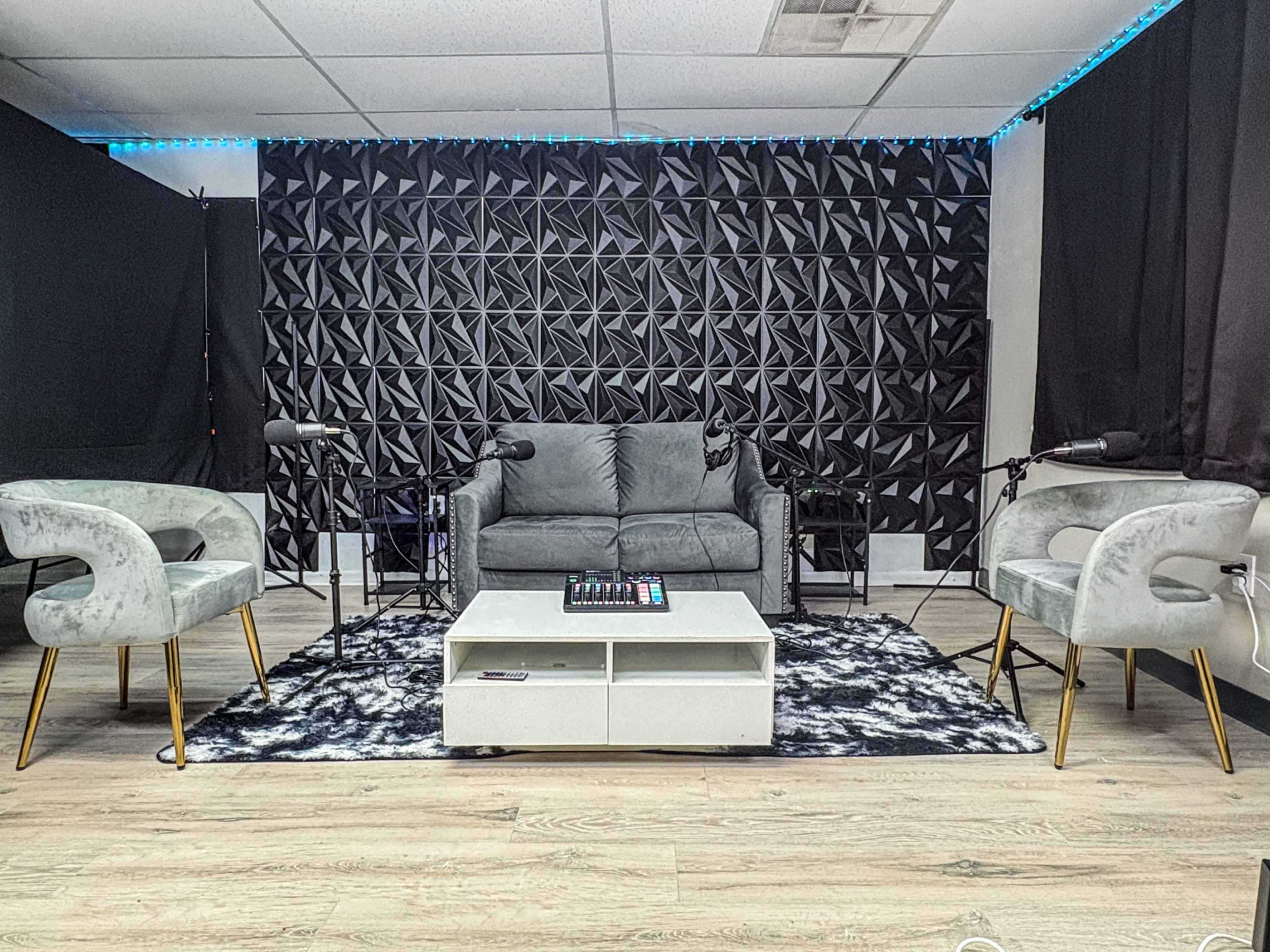 The image shows a modern recording studio setup featuring a gray couch, four chairs, a coffee table, and multiple microphones on stands against a textured black wall.
