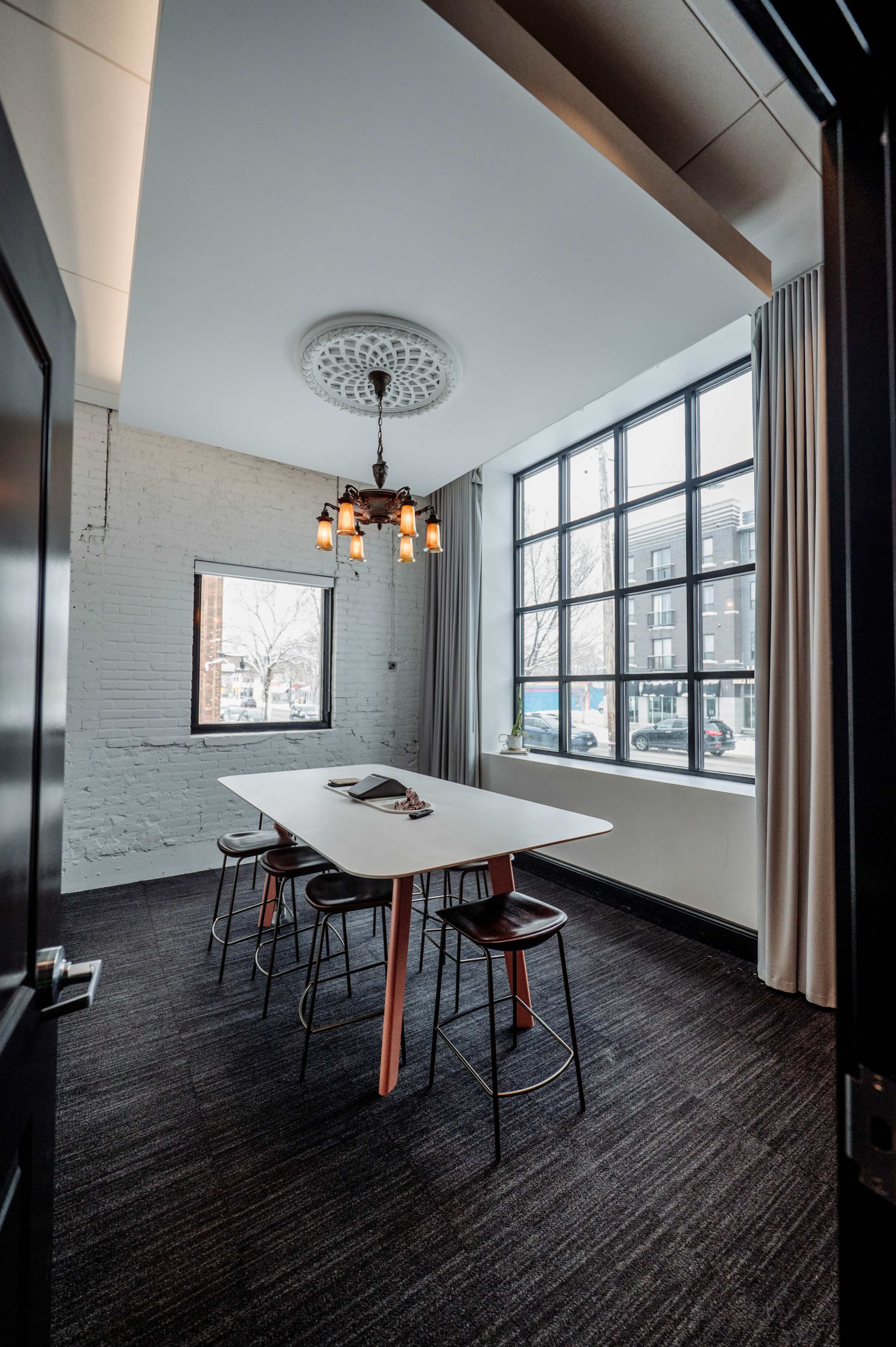 A modern meeting room features a large white table surrounded by black stools, with a decorative chandelier and large windows bringing in natural light.