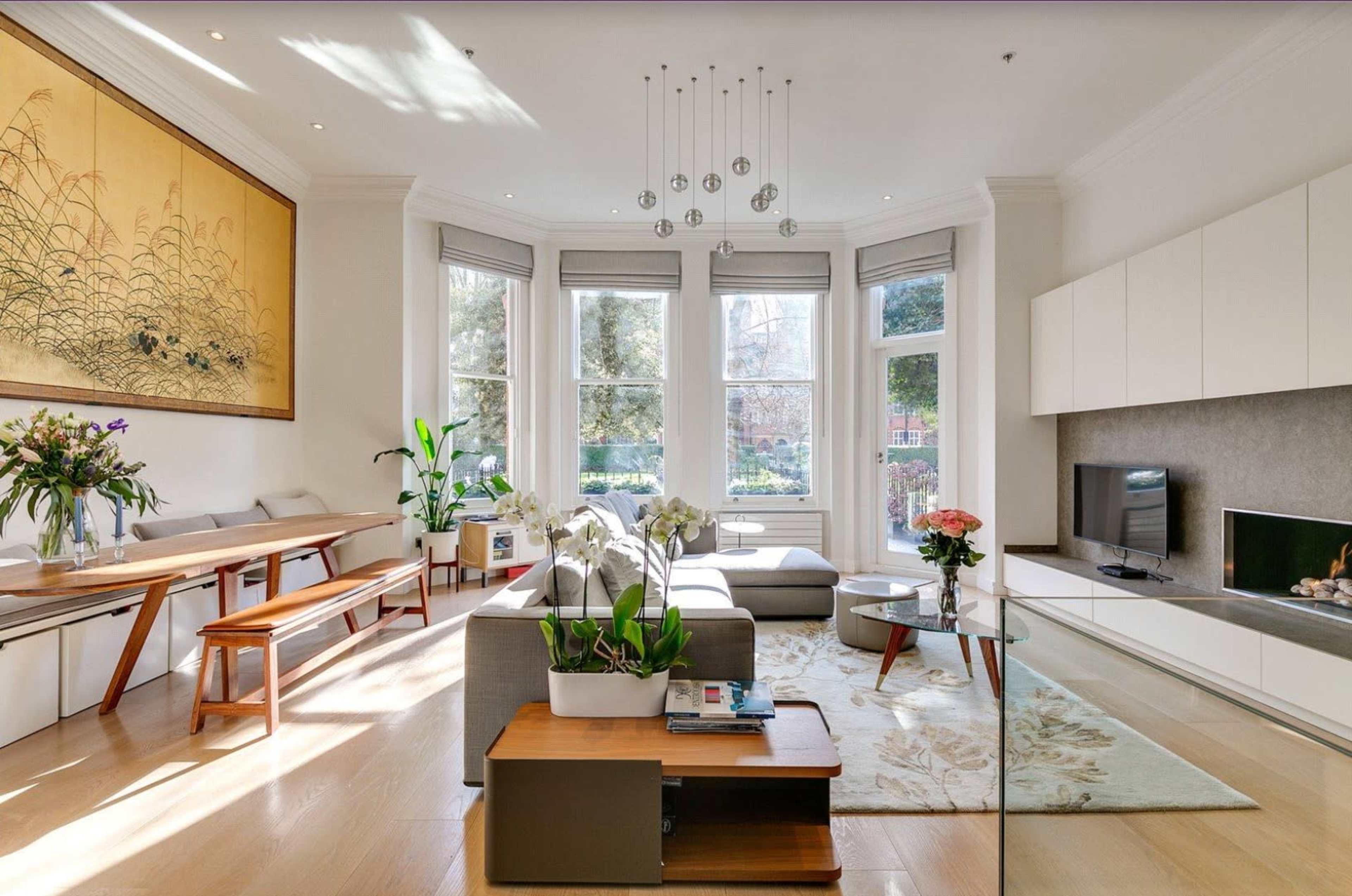 A bright, modern living room features a large artwork, a sectional sofa, a coffee table, and a window with views of greenery outside.