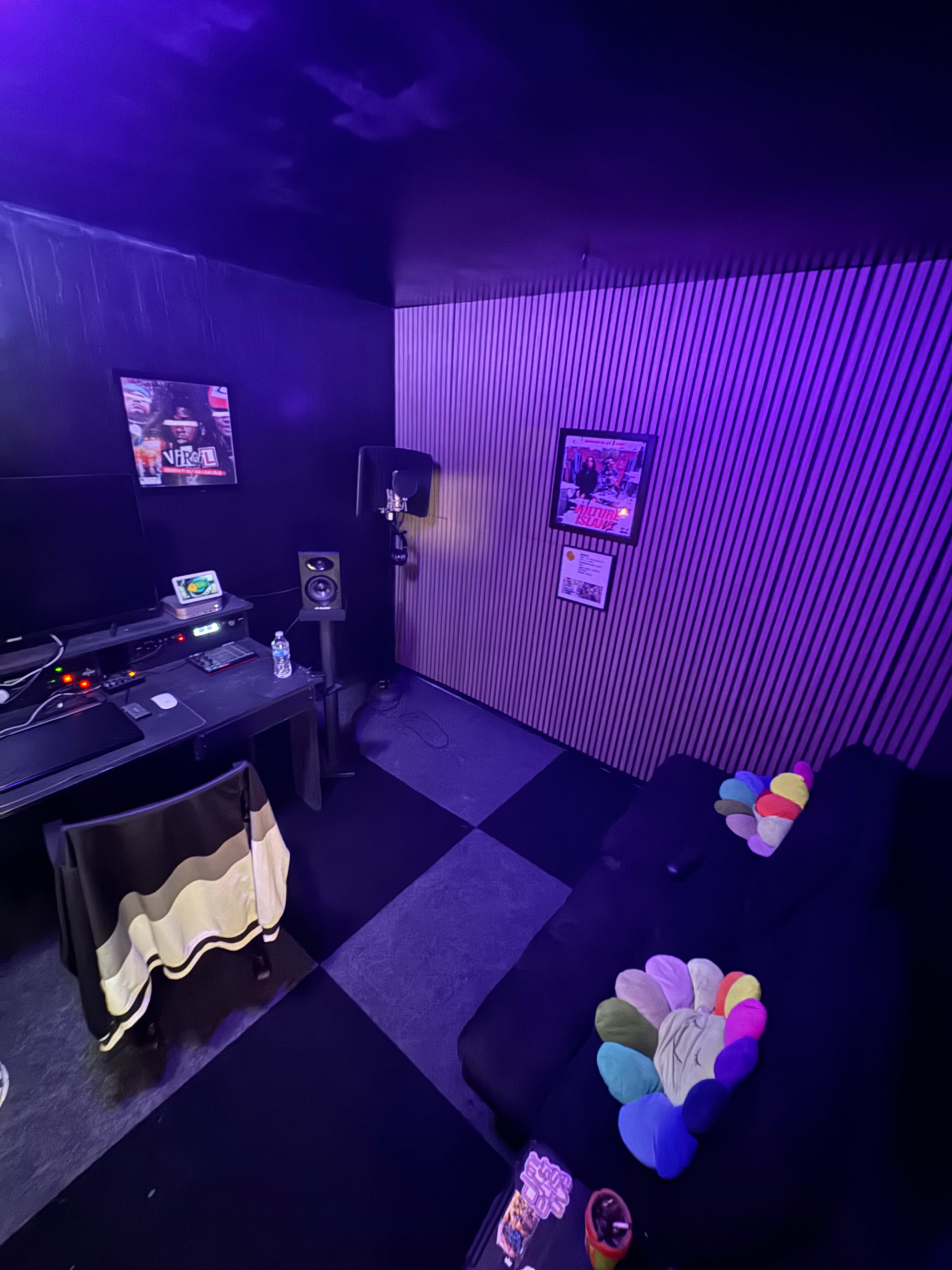 The image shows a small, dimly lit music studio with a digital audio workstation setup on a desk, a microphone on a stand, and a couch with colorful cushions along one side.