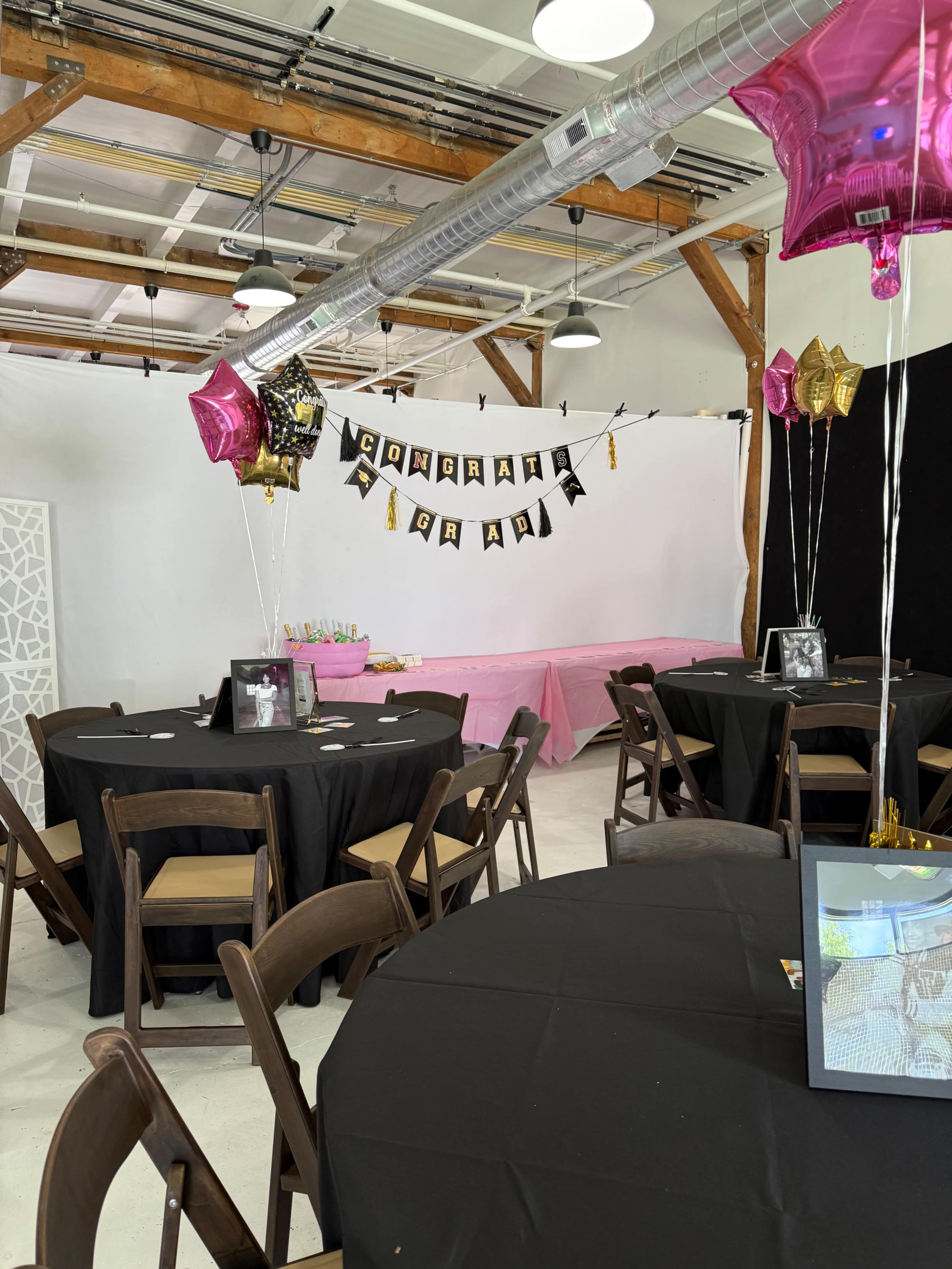 The image shows a decorated event space with black and pink table settings, featuring a "Congratulations" banner and festive balloons.