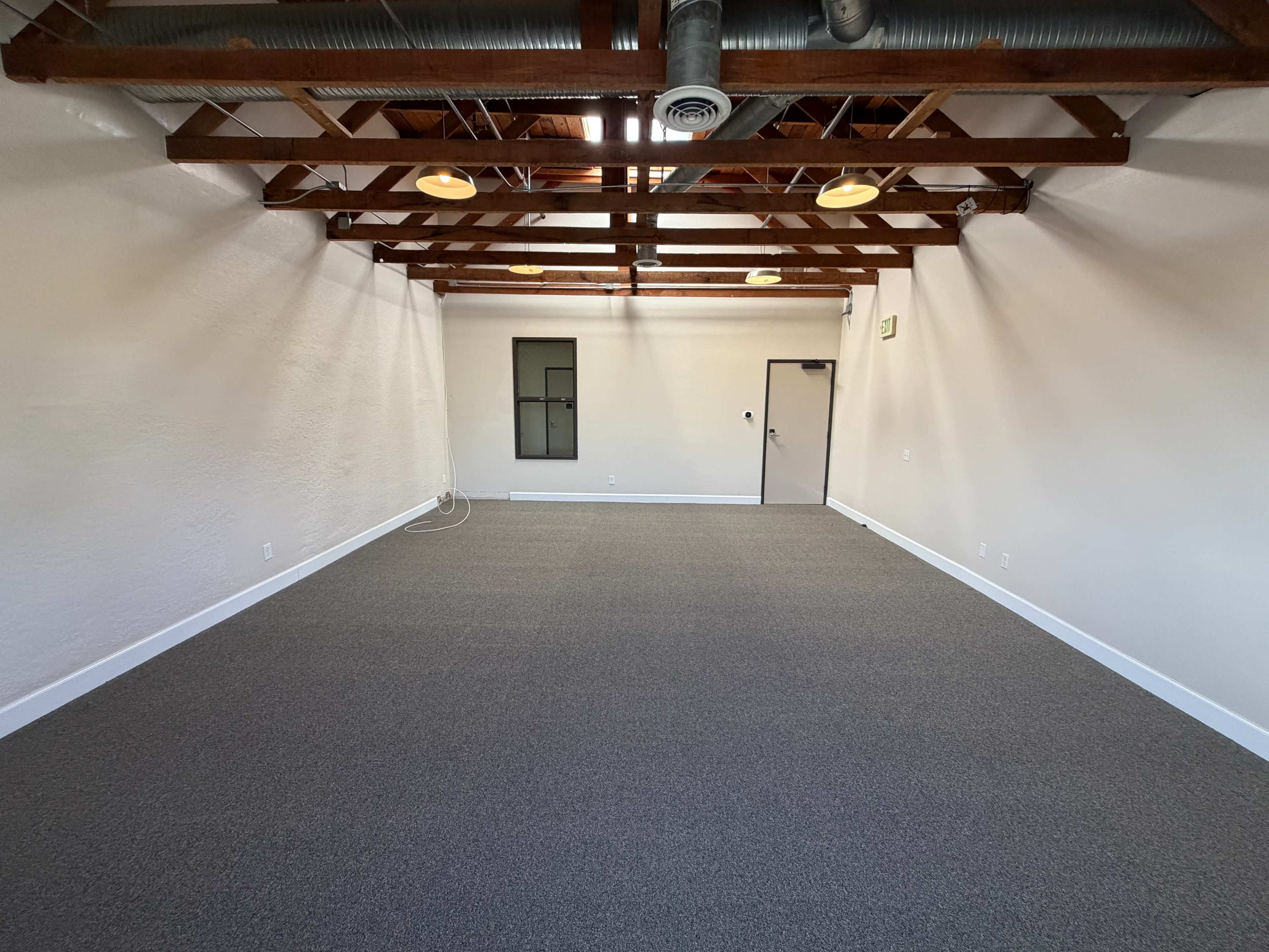 The image shows an empty room with a carpeted floor, exposed wooden beams, and a door on one side leading to another space.