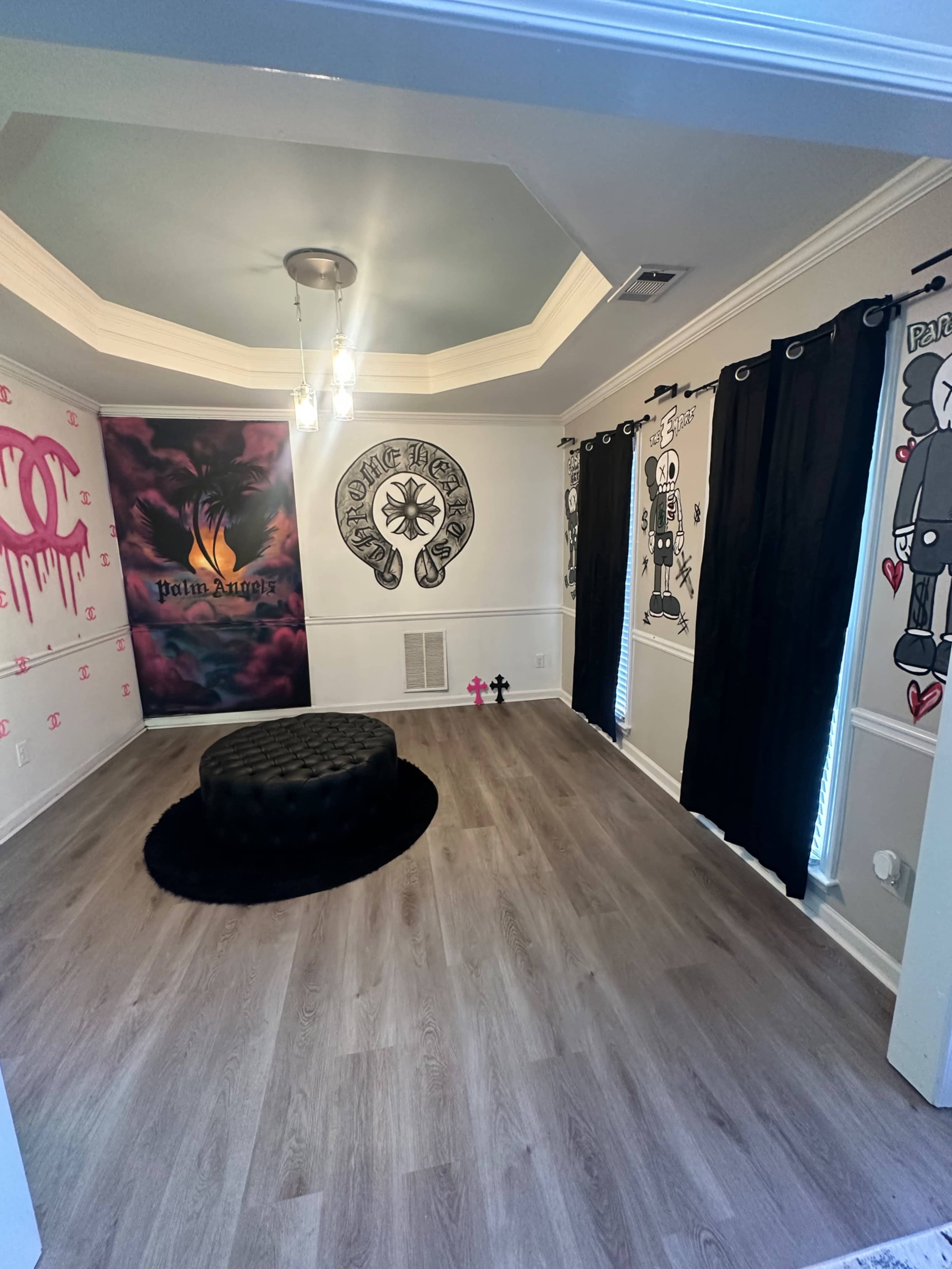 The room features a circular chandelier, dark curtains, and decorated walls with artwork, along with a black ottoman in the center.