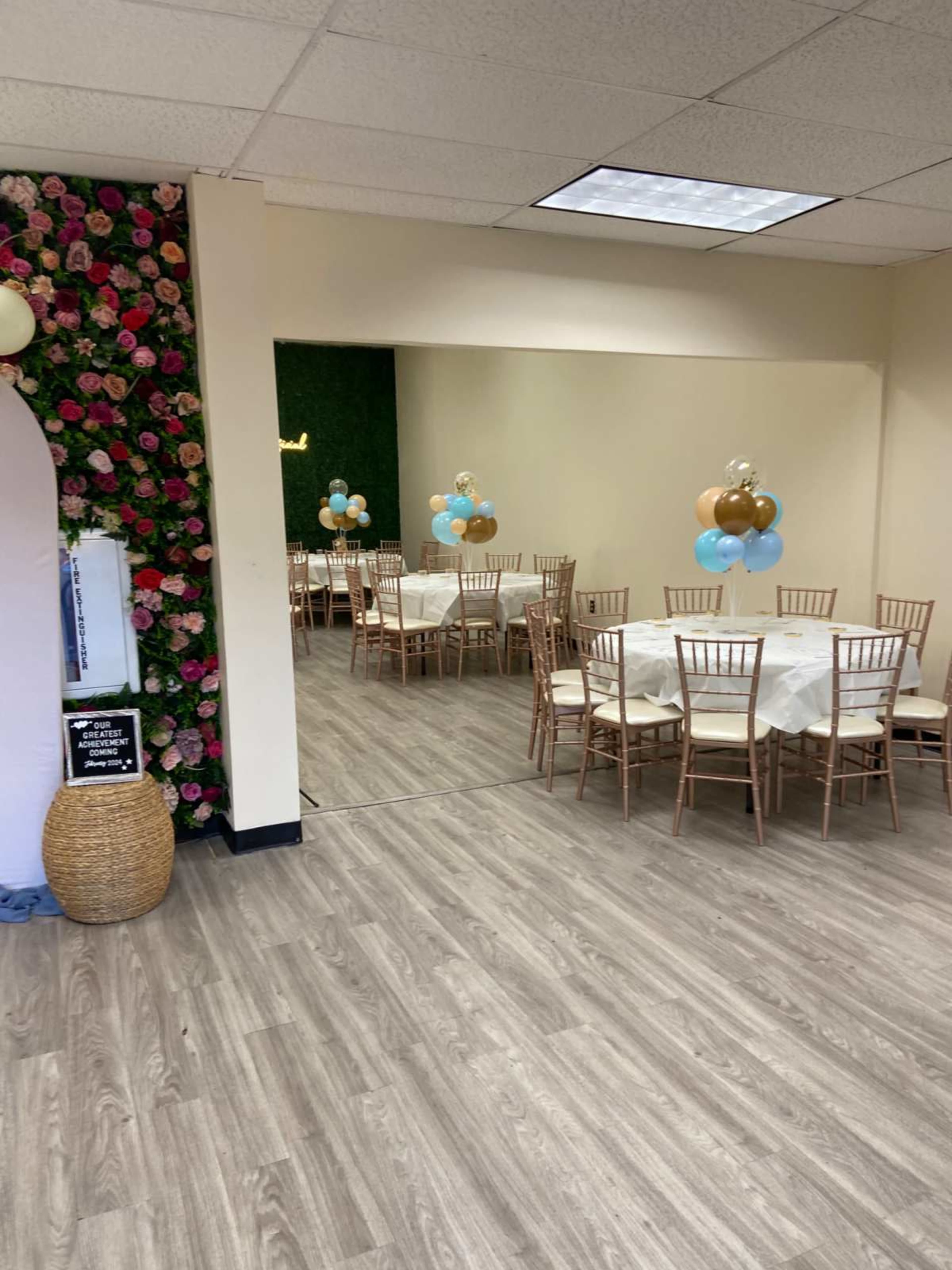 A decorated event space with multiple tables set with white tablecloths and chairs, along with balloon arrangements in blue and gold.