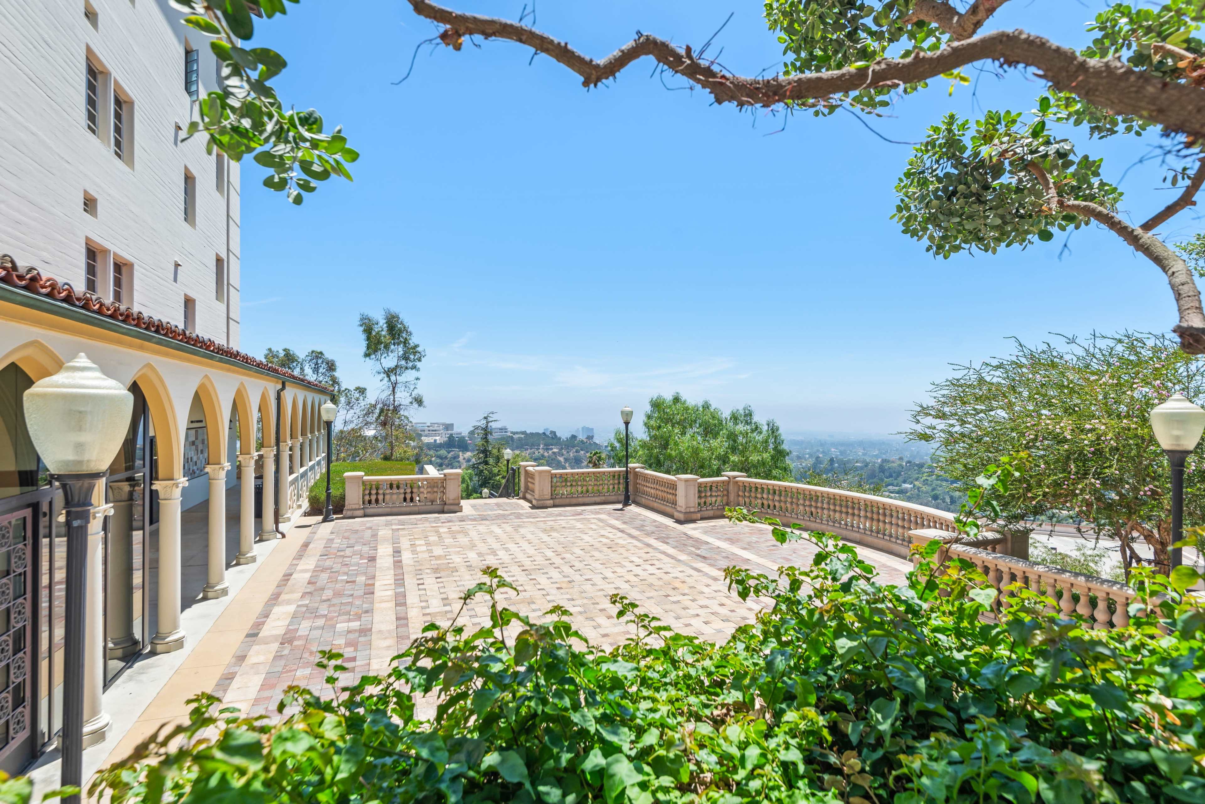The image shows a landscaped outdoor area with a paved patio, arched architecture, and a view of distant hills in clear weather.