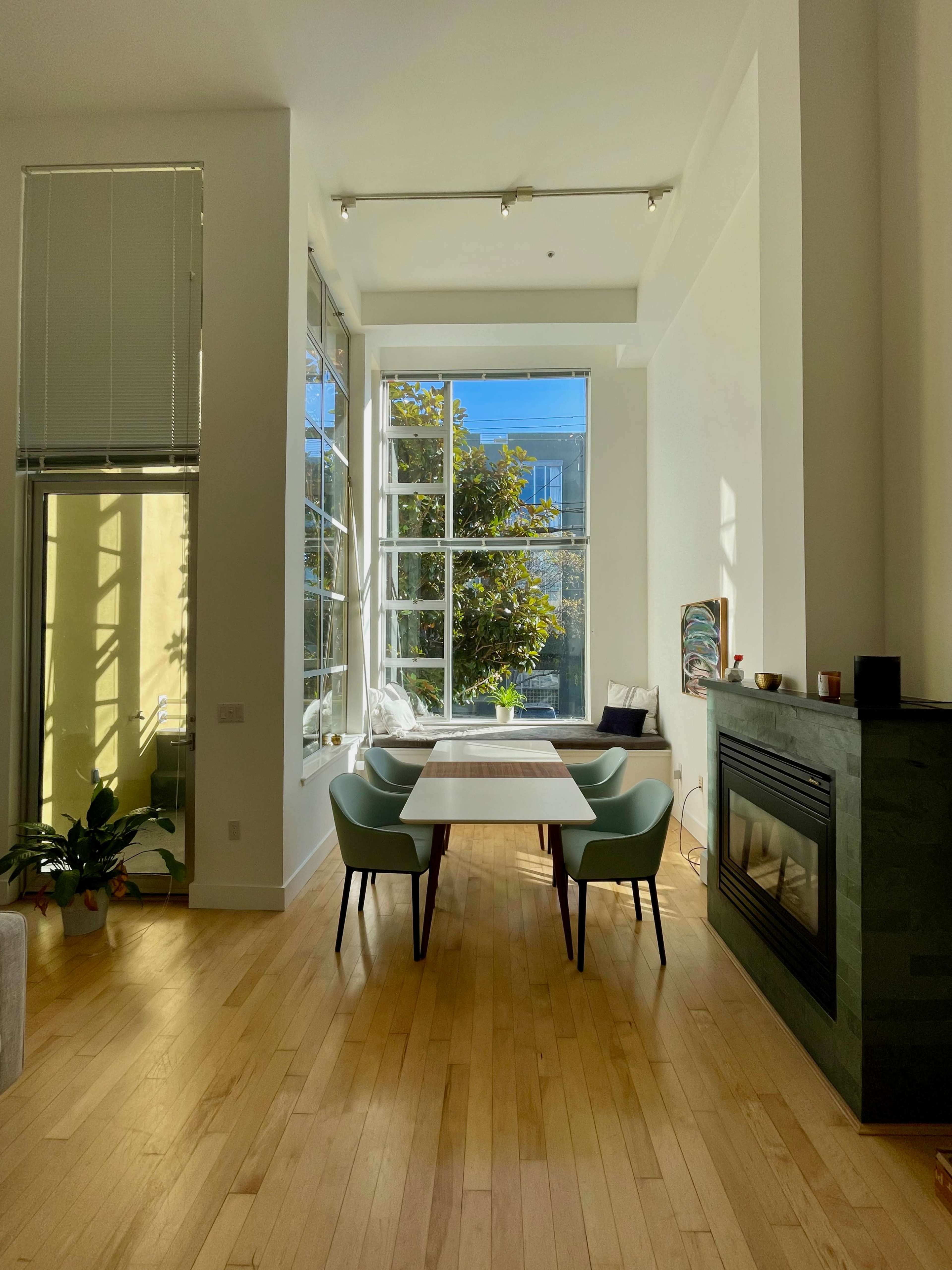 A bright, modern dining area features a wooden table surrounded by two chairs, with large windows letting in natural light.