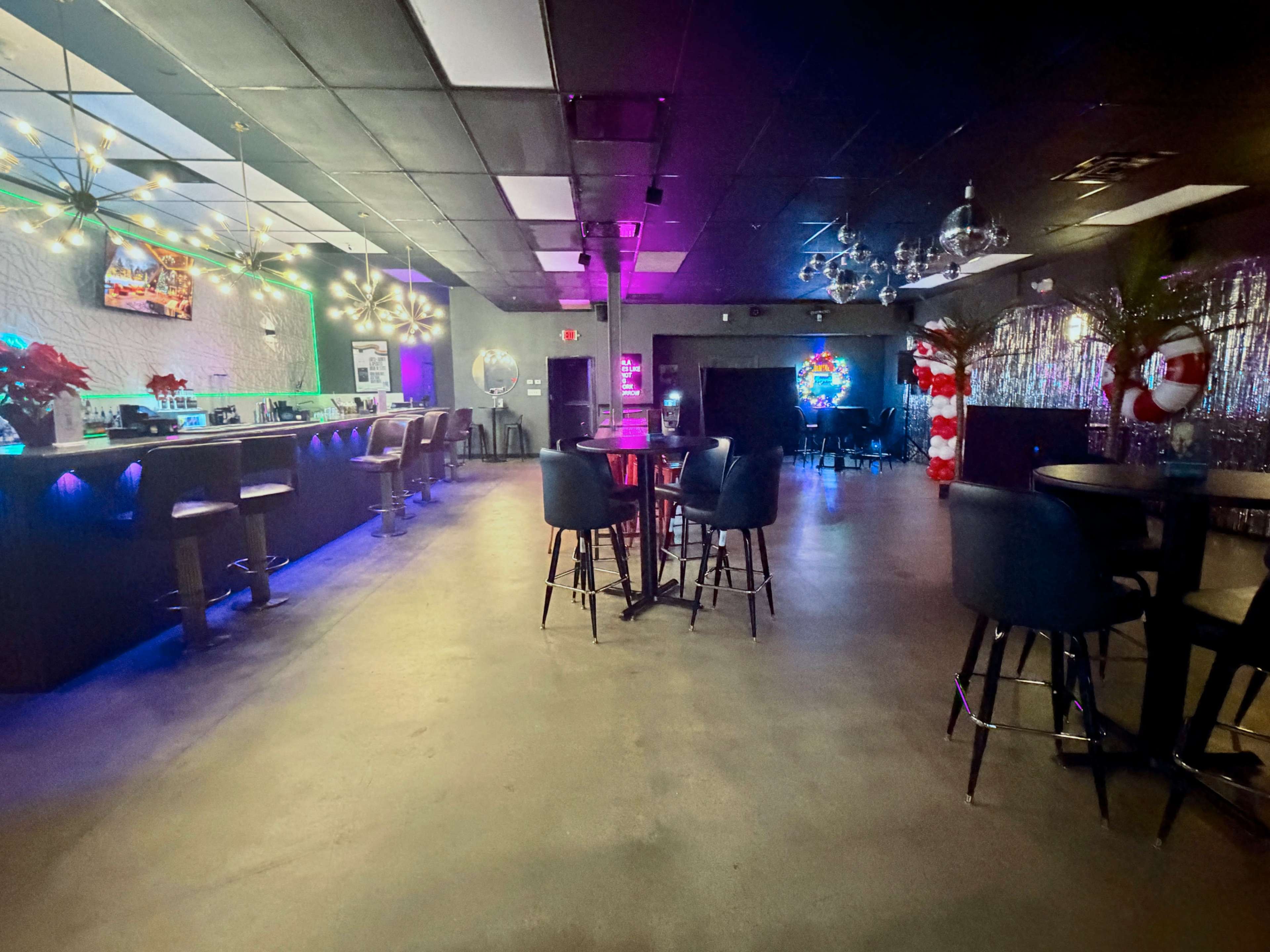 A dimly lit event space features a bar area with stools, colorful lighting, and festive decorations.