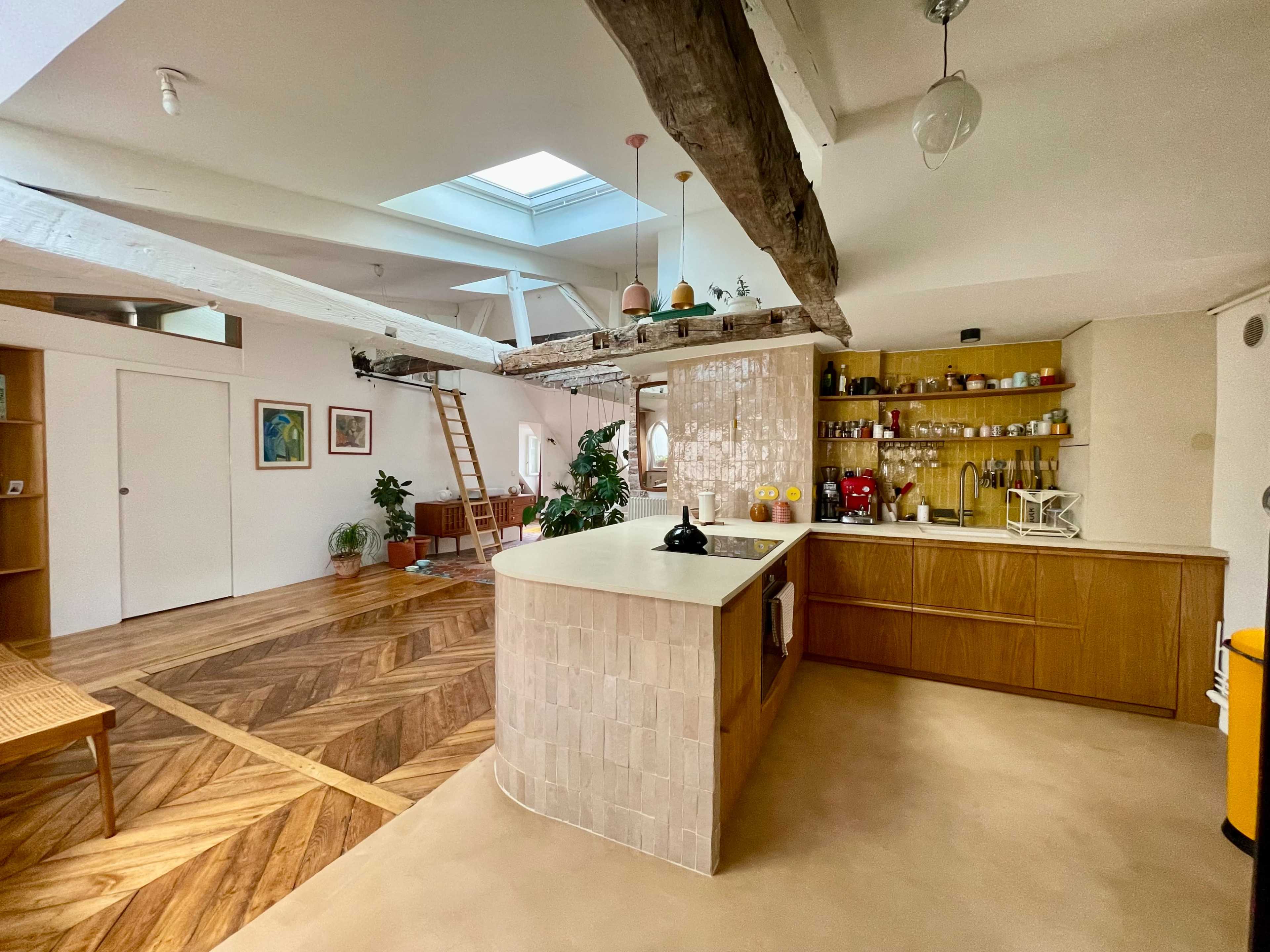 A modern, open-concept kitchen and living area features a large island, wooden cabinetry, and a staircase leading to a loft space, with natural light coming from a skylight above.