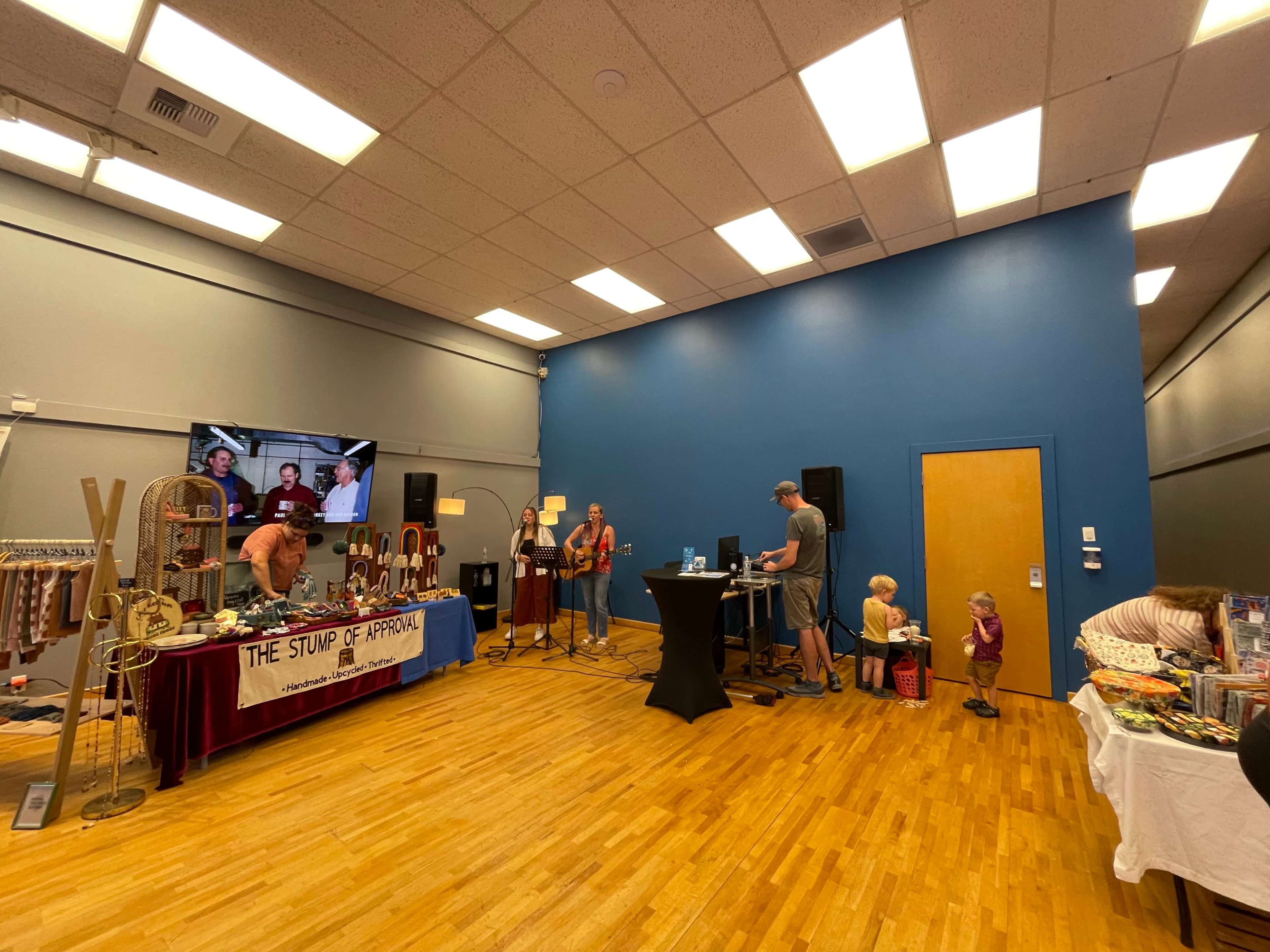 A spacious indoor venue with vendors displaying goods on tables, a stage area with a man using a microphone, and a child observing nearby.