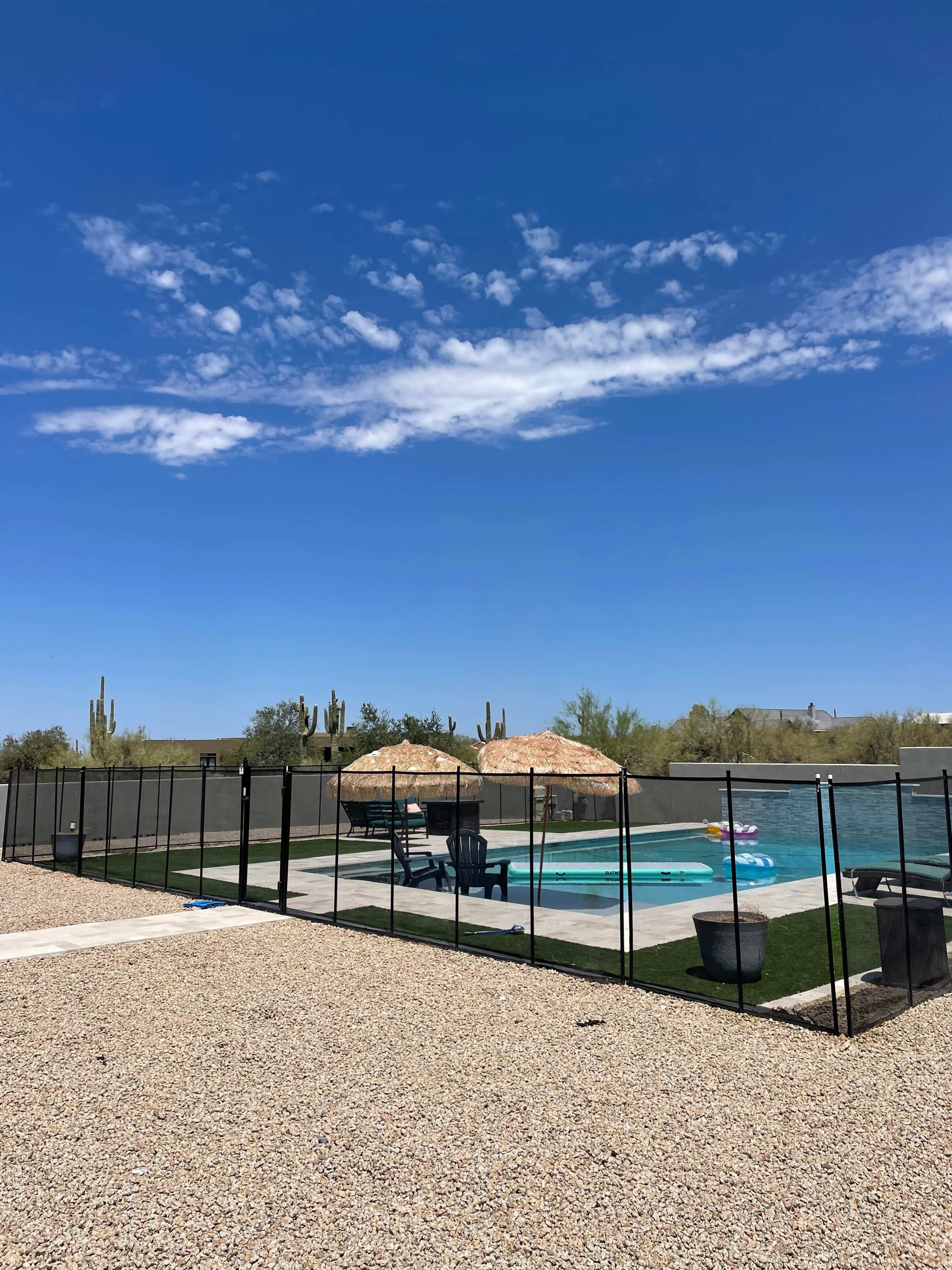 A swimming pool with two thatched umbrellas and lounge chairs is enclosed by a black safety fence against a backdrop of a clear blue sky and desert landscape.