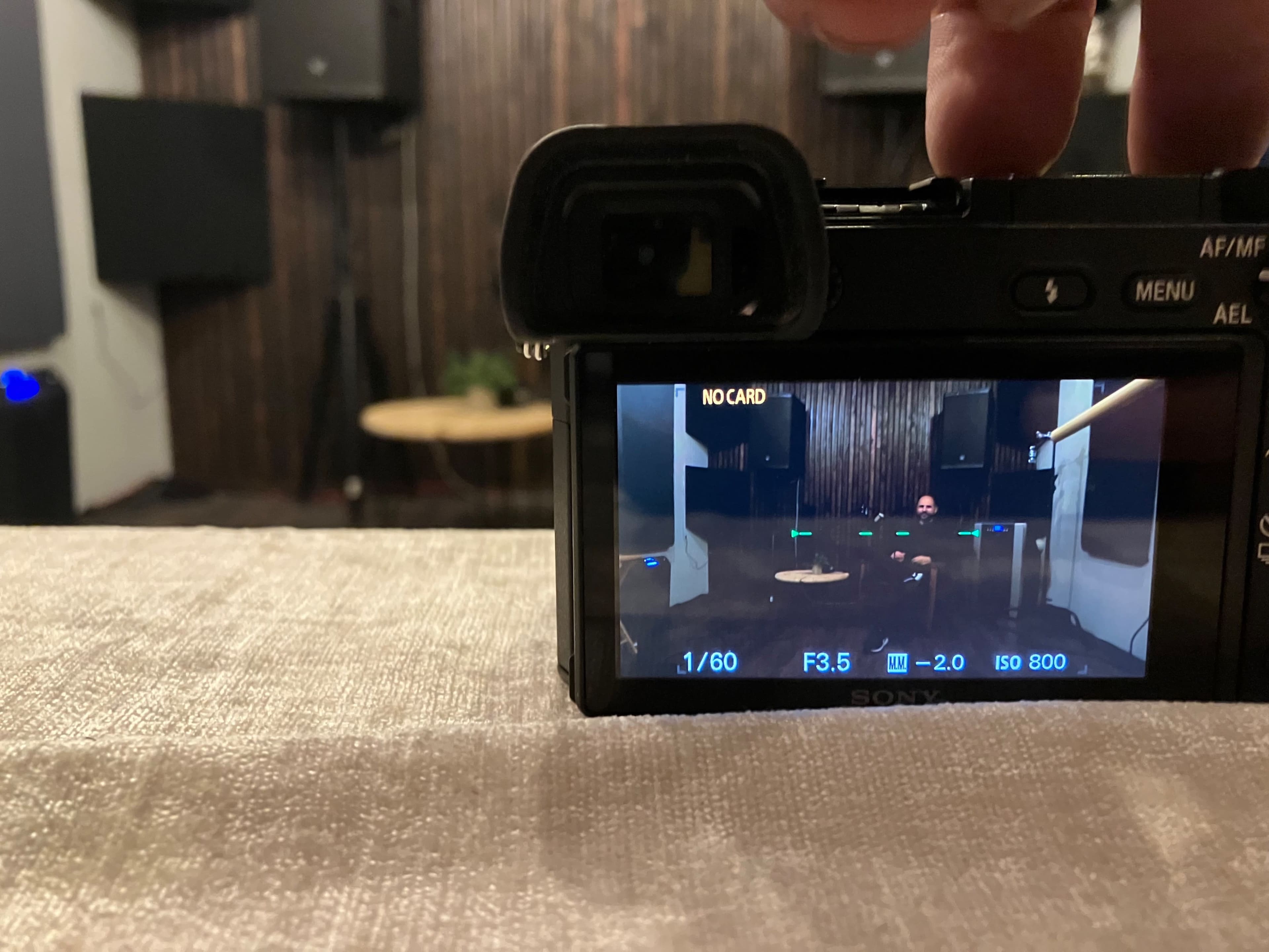 A camera on a table captures a view of a person seated in a dimly lit room with wooden paneling and sound equipment.