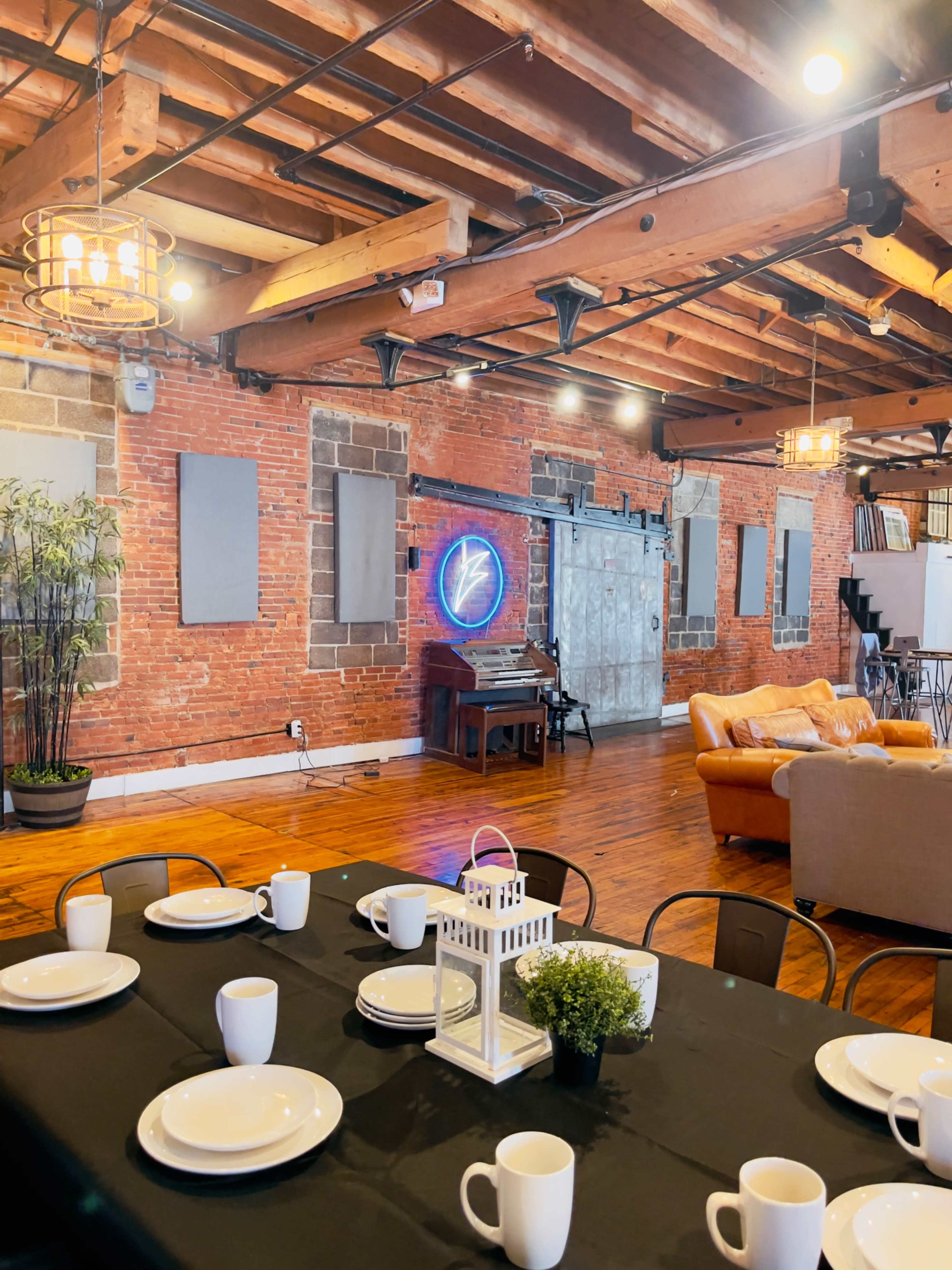 The image shows a spacious, rustic interior with exposed brick walls, wooden beams, and a table set for dining, featuring plates, cups, and a small plant centerpiece.
