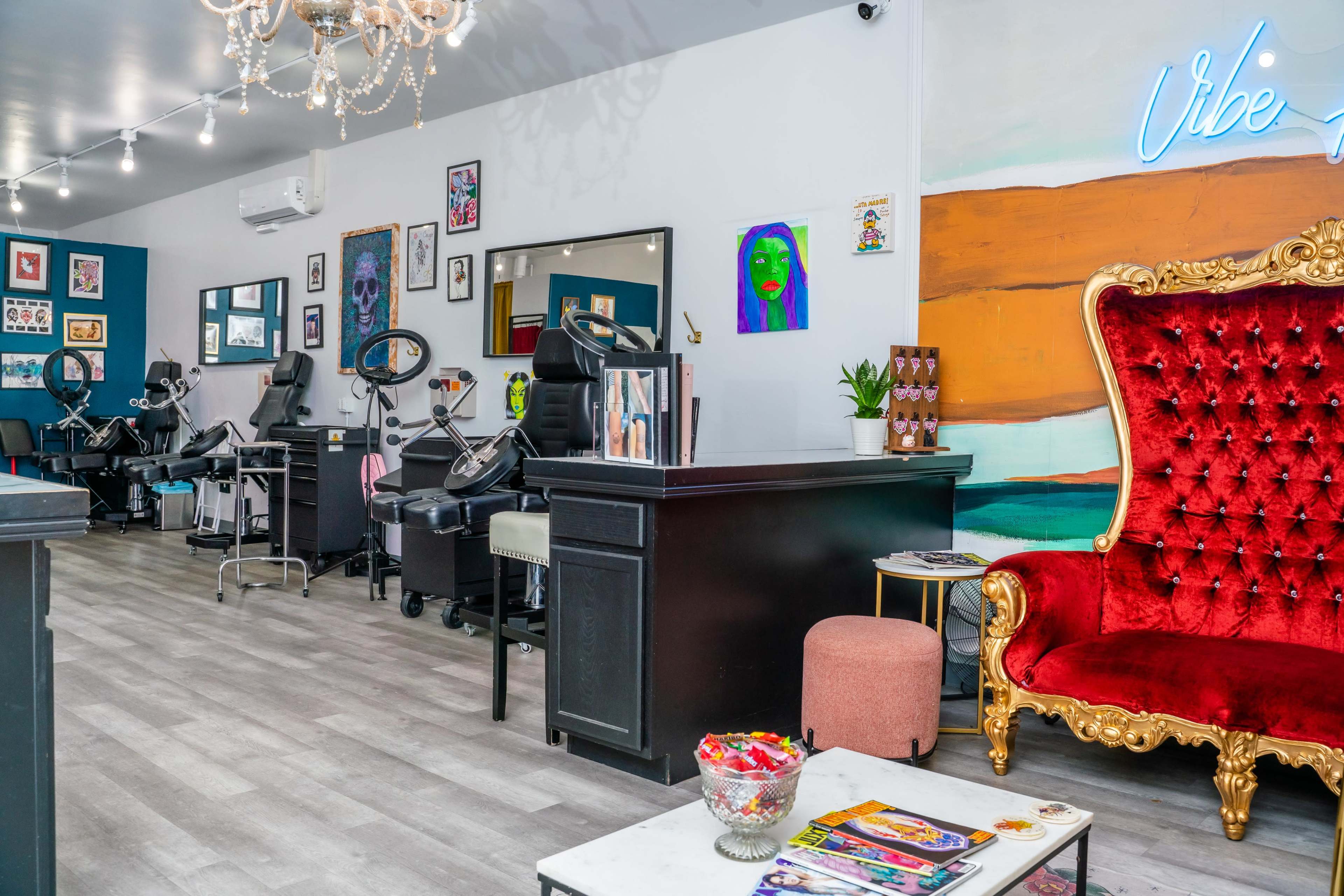 The image shows a tattoo parlor with several tattoo stations, a decorative red chair, and various artworks displayed on the walls.
