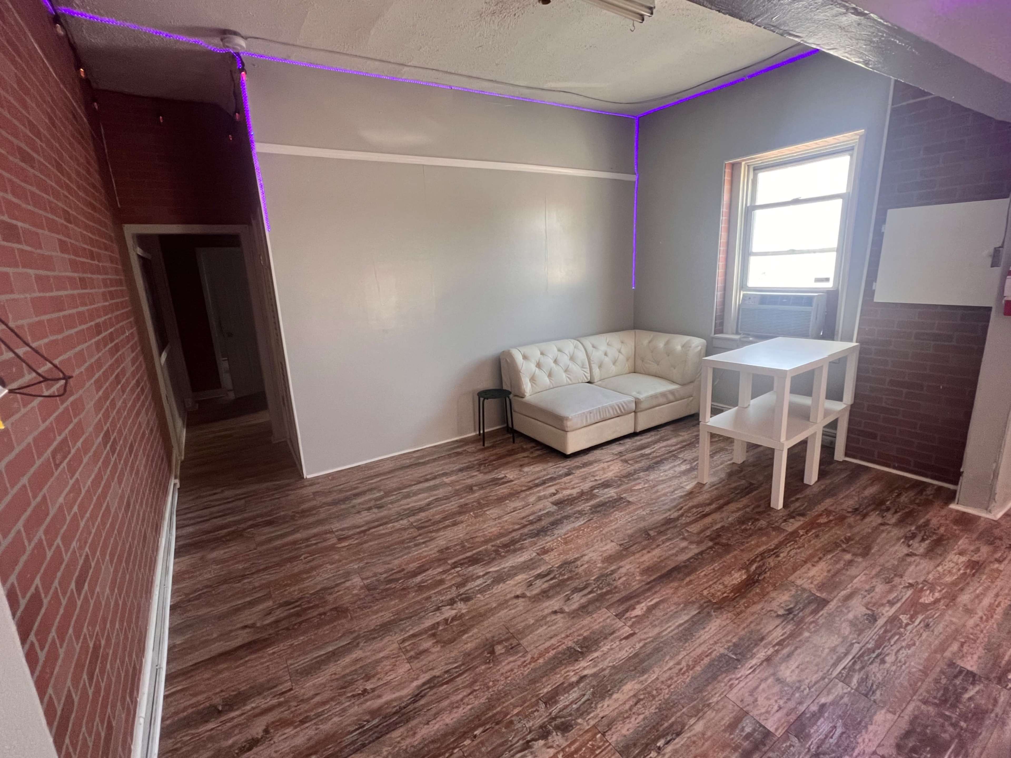 The image shows an interior room with a white couch, a small table, and a window, featuring exposed brick walls and wooden flooring.