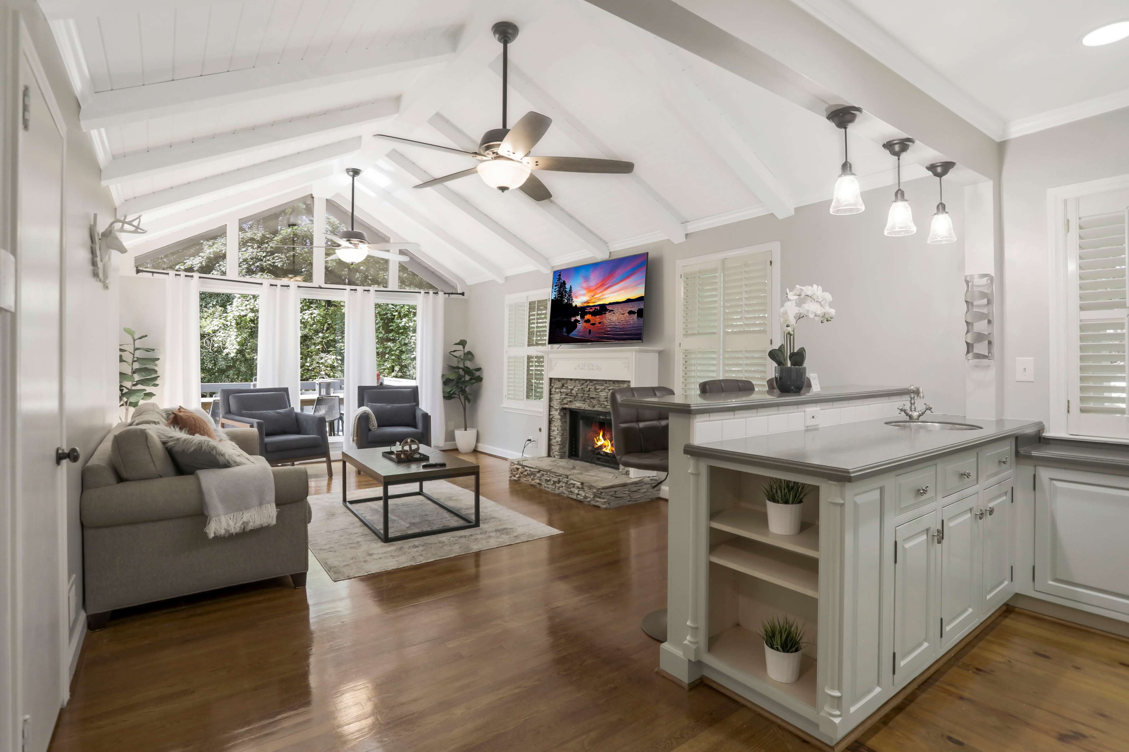 A spacious living area features vaulted ceilings, a stone fireplace, large windows overlooking a patio, and modern furnishings arranged around a central coffee table.