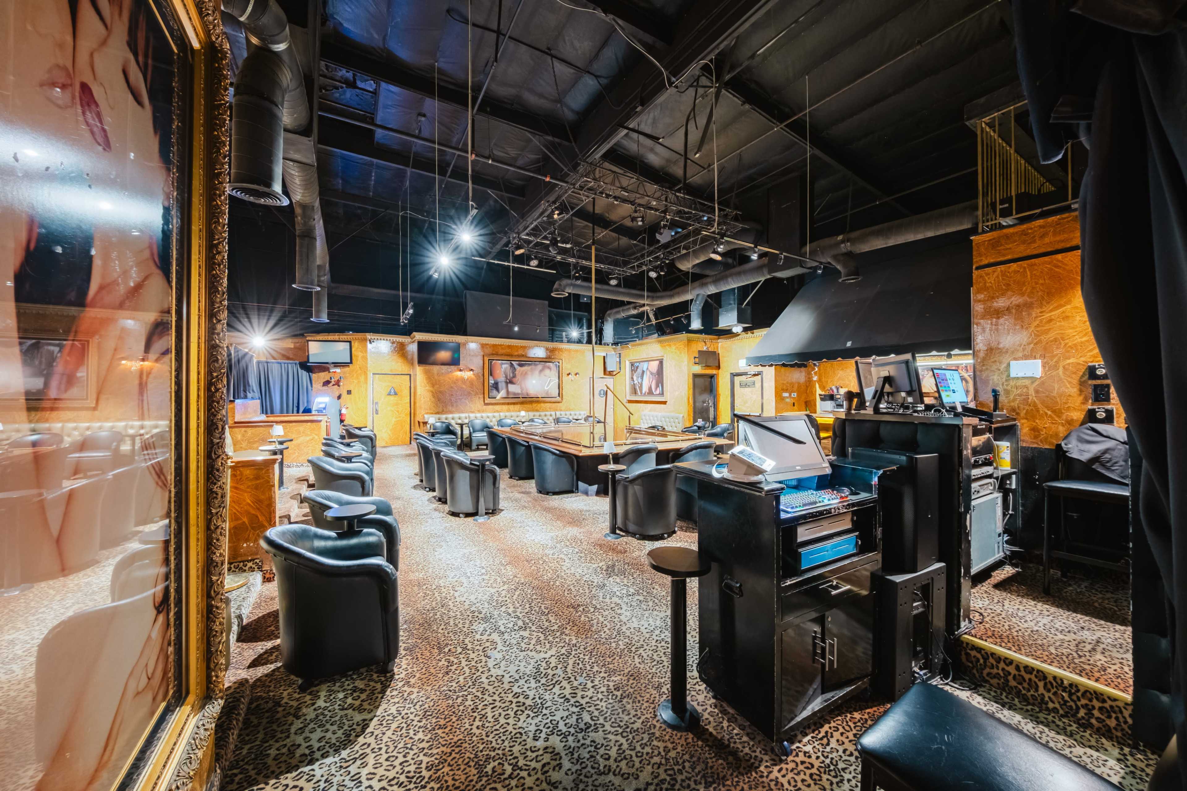 The image shows the interior of a dimly lit lounge or bar featuring black leather chairs, a poker table, and machines along the walls.