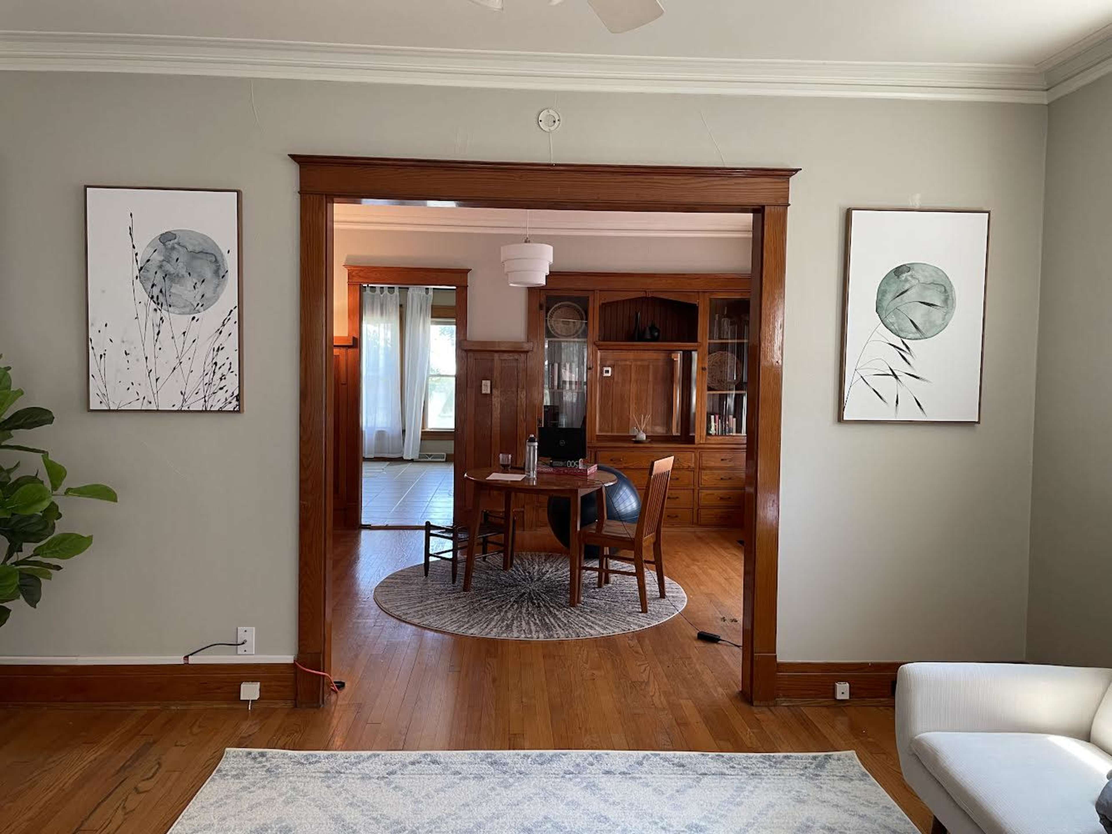 An interior setting with two framed artworks on a light gray wall, leading to a dining area furnished with a round table and four chairs.