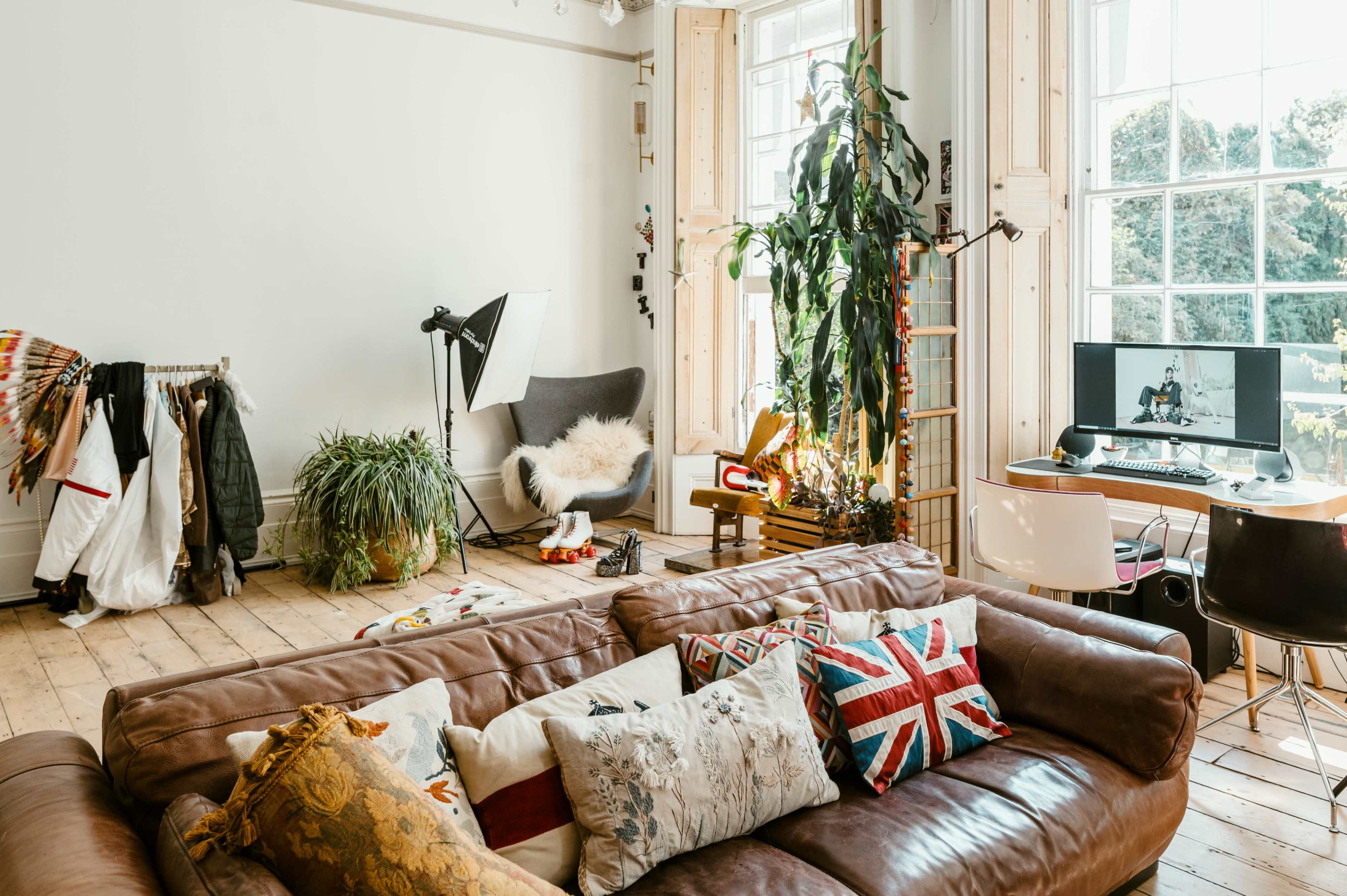 The image shows a cozy living room with a brown leather sofa adorned with colorful cushions, a large window letting in natural light, and a workspace featuring a computer and a houseplant.
