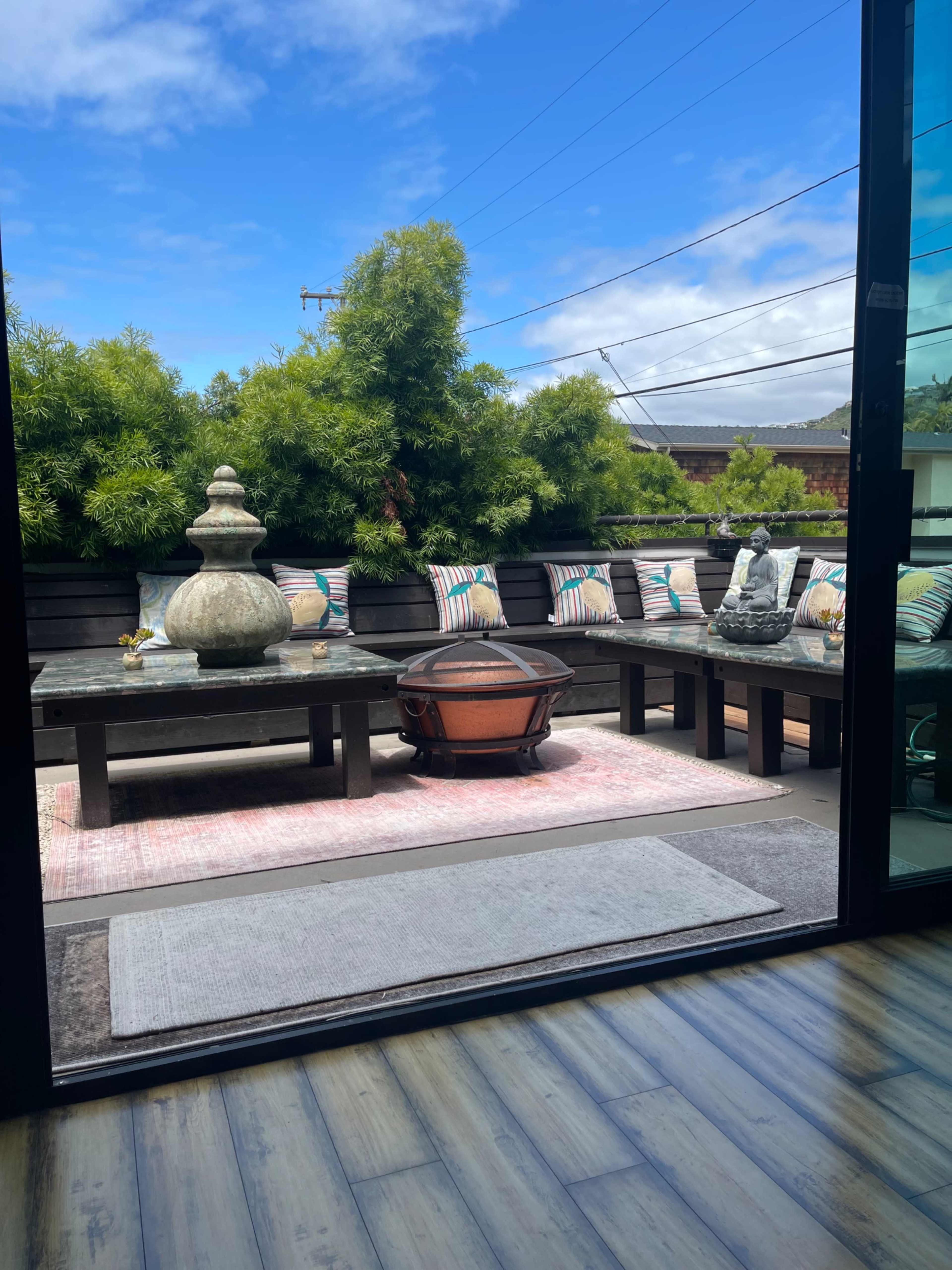 A patio with a circular fire pit, a sectional seating area adorned with cushions, and potted greenery under a clear blue sky.