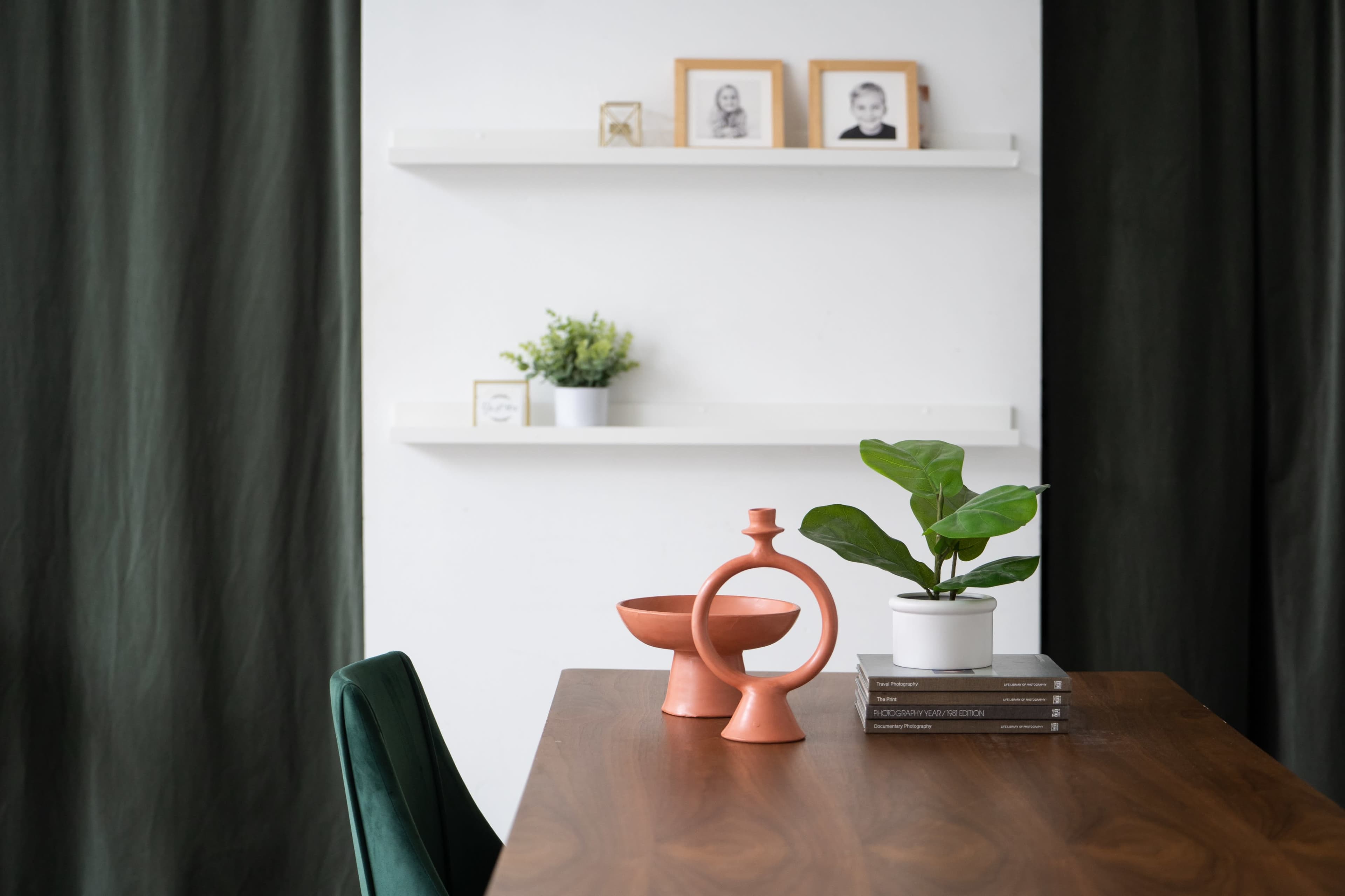 A wooden table is set with a terracotta vase and bowl, alongside a small potted plant and neatly stacked books, with framed photos on a shelf in the background.