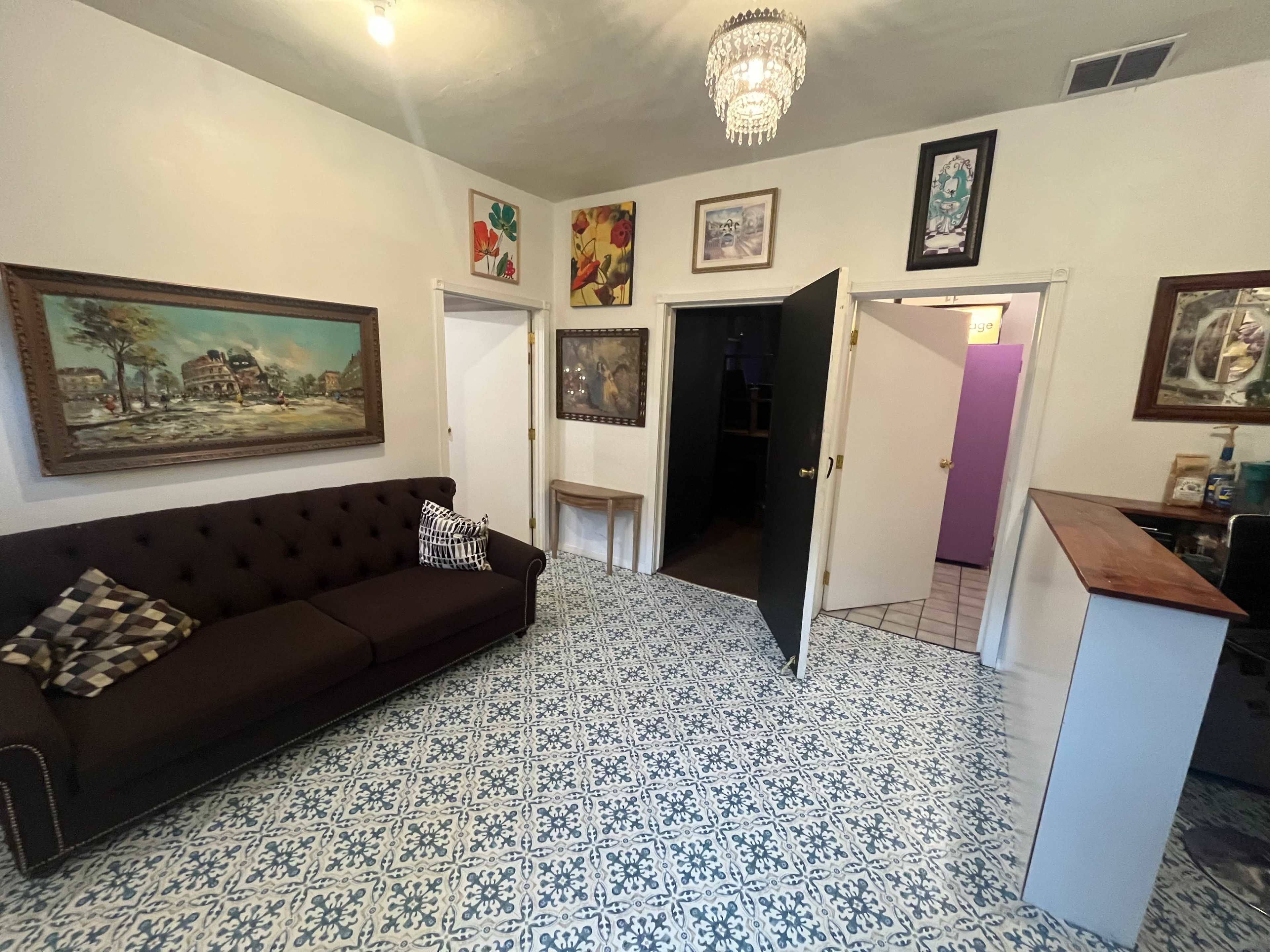 The image shows a small room with a dark brown sofa, decorative artwork on the walls, and two doors leading to adjacent spaces.