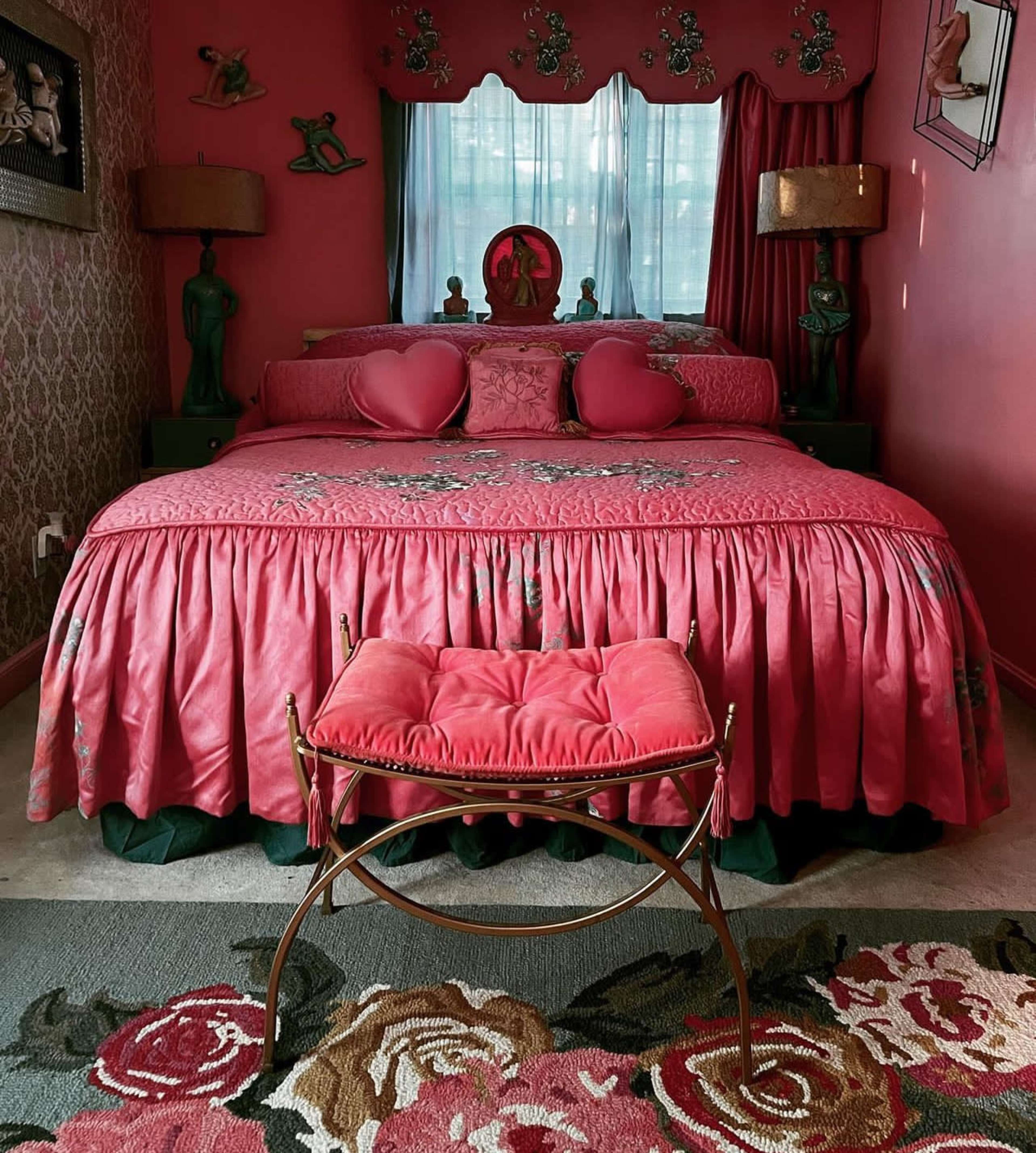 The room features a large pink bed with decorative pillows and a matching ruffled bed skirt, flanked by two lamps, alongside a metal stool and a floral rug.
