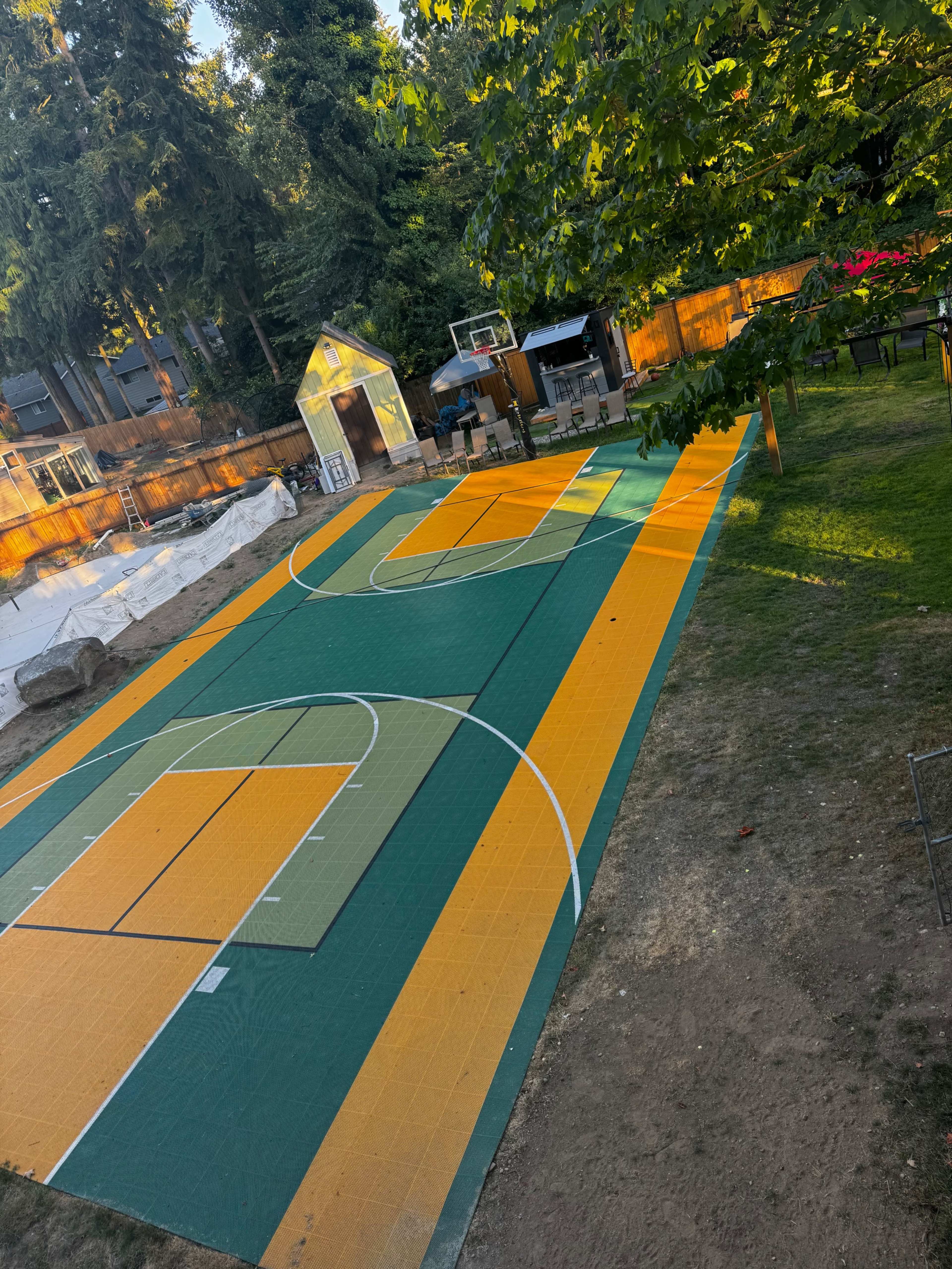The image shows a sports court with vibrant green and orange tiles, set in a backyard surrounded by trees and a storage shed.