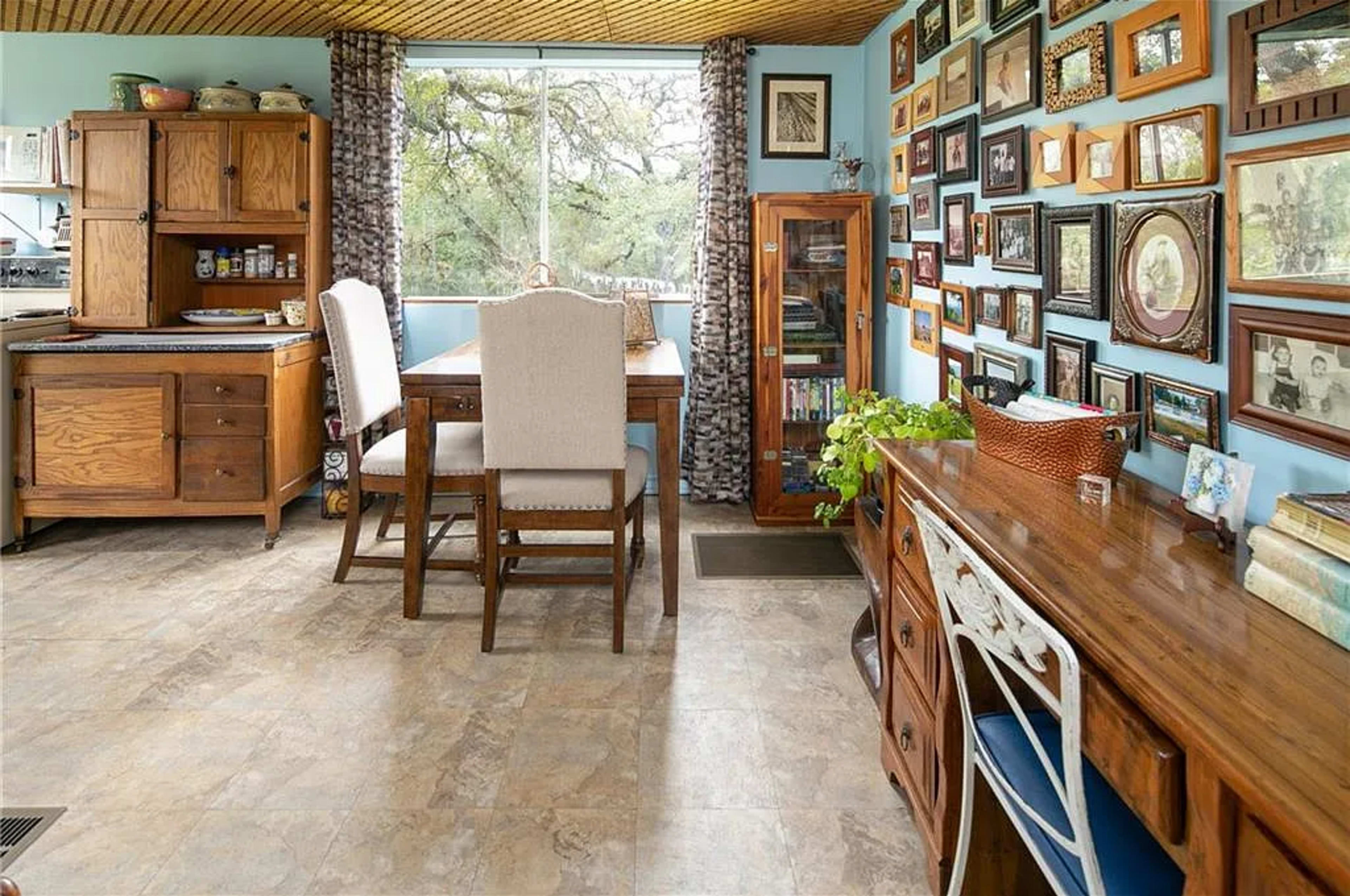 The image depicts a cozy kitchen and dining area featuring wooden furniture, including a table with chairs and a sideboard, surrounded by a wall adorned with various framed photographs.