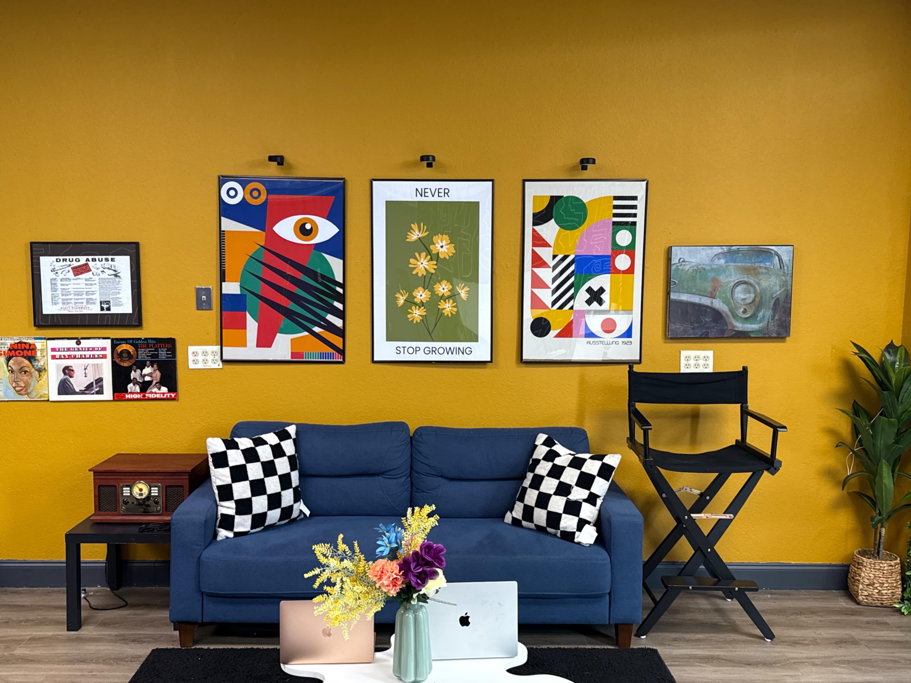 The image shows a cozy interior with a blue sofa, a vibrant orange wall, decorative artwork hanging above the sofa, and a small table with a laptop and a floral arrangement.