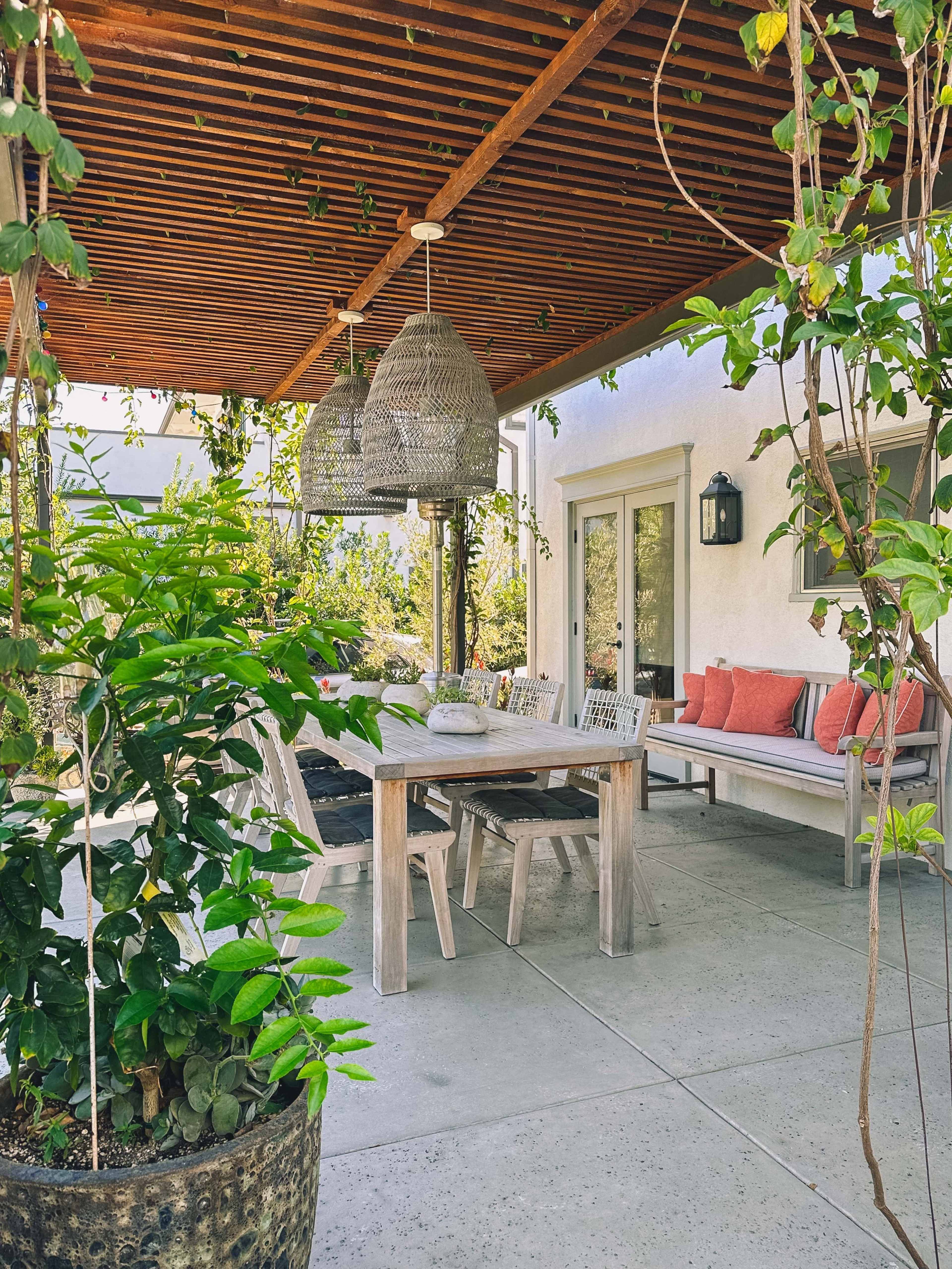 A covered outdoor patio with a wooden dining table and chairs, surrounded by greenery and decorative hanging lights.
