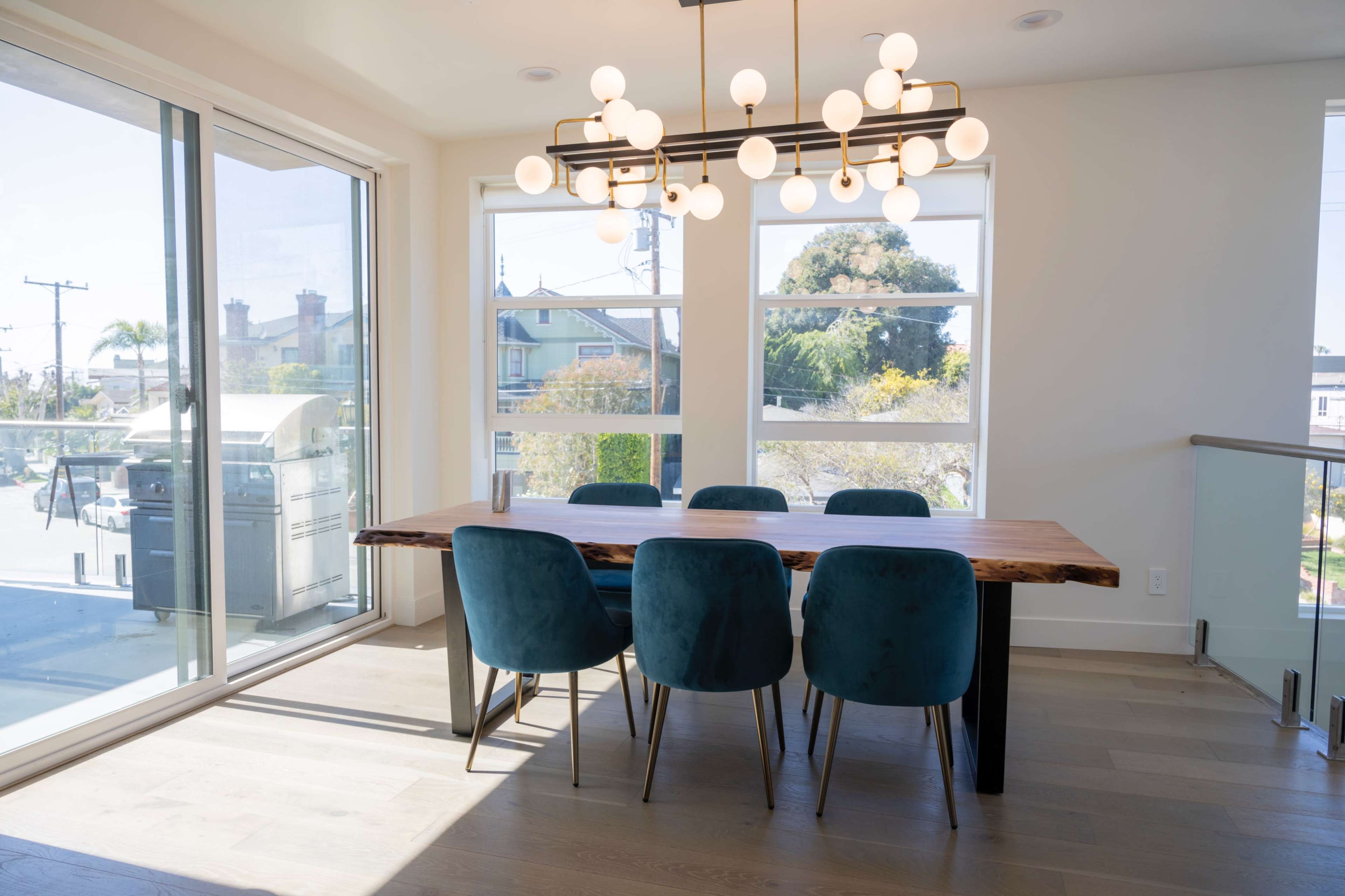 A modern dining area features a wooden table surrounded by six blue chairs, illuminated by a stylish chandelier, with large windows providing natural light and a view of the outdoors.
