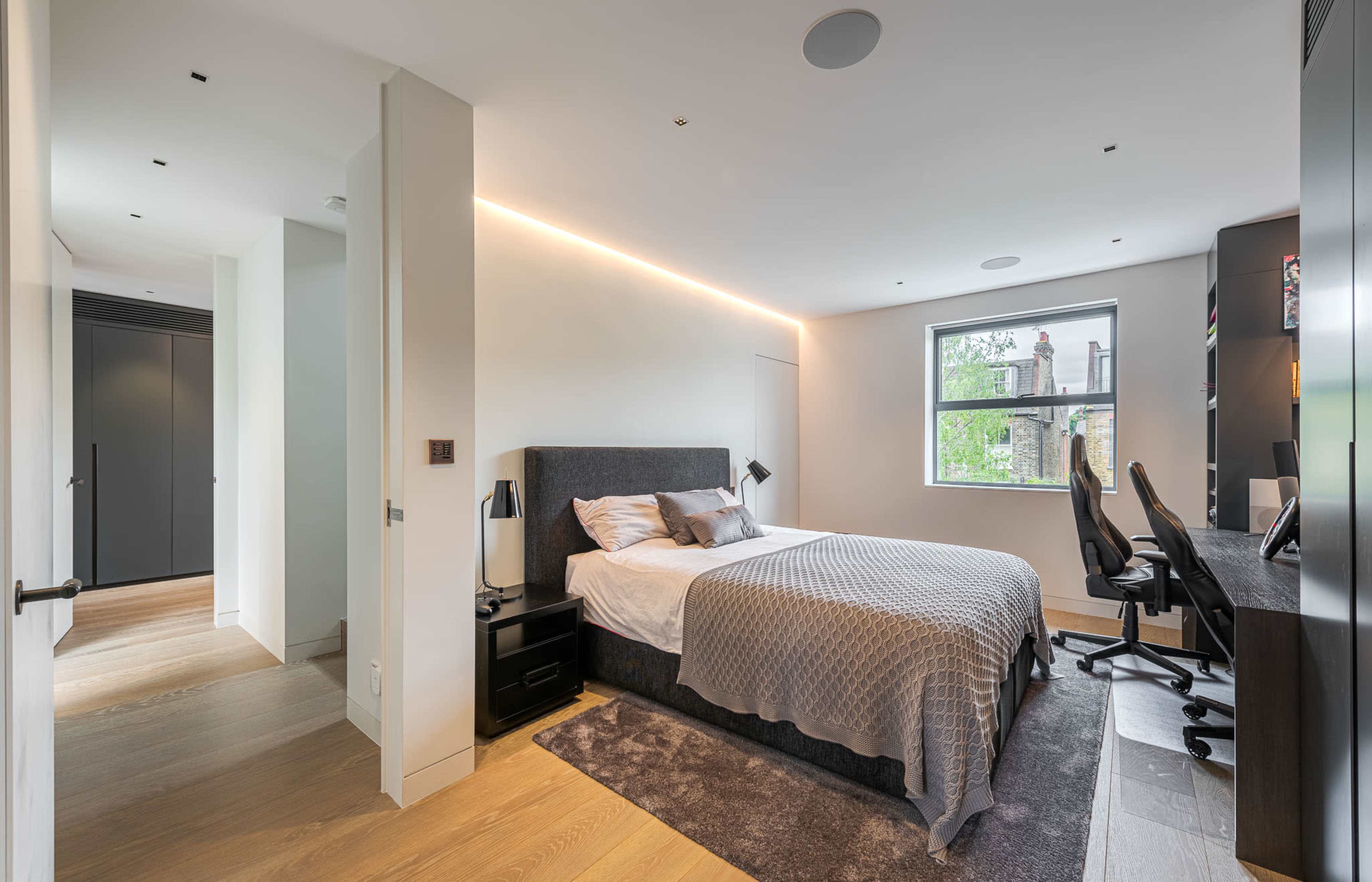 A modern bedroom with a bed, a desk with chairs, and a window offering natural light, complemented by light-colored walls and wooden flooring.
