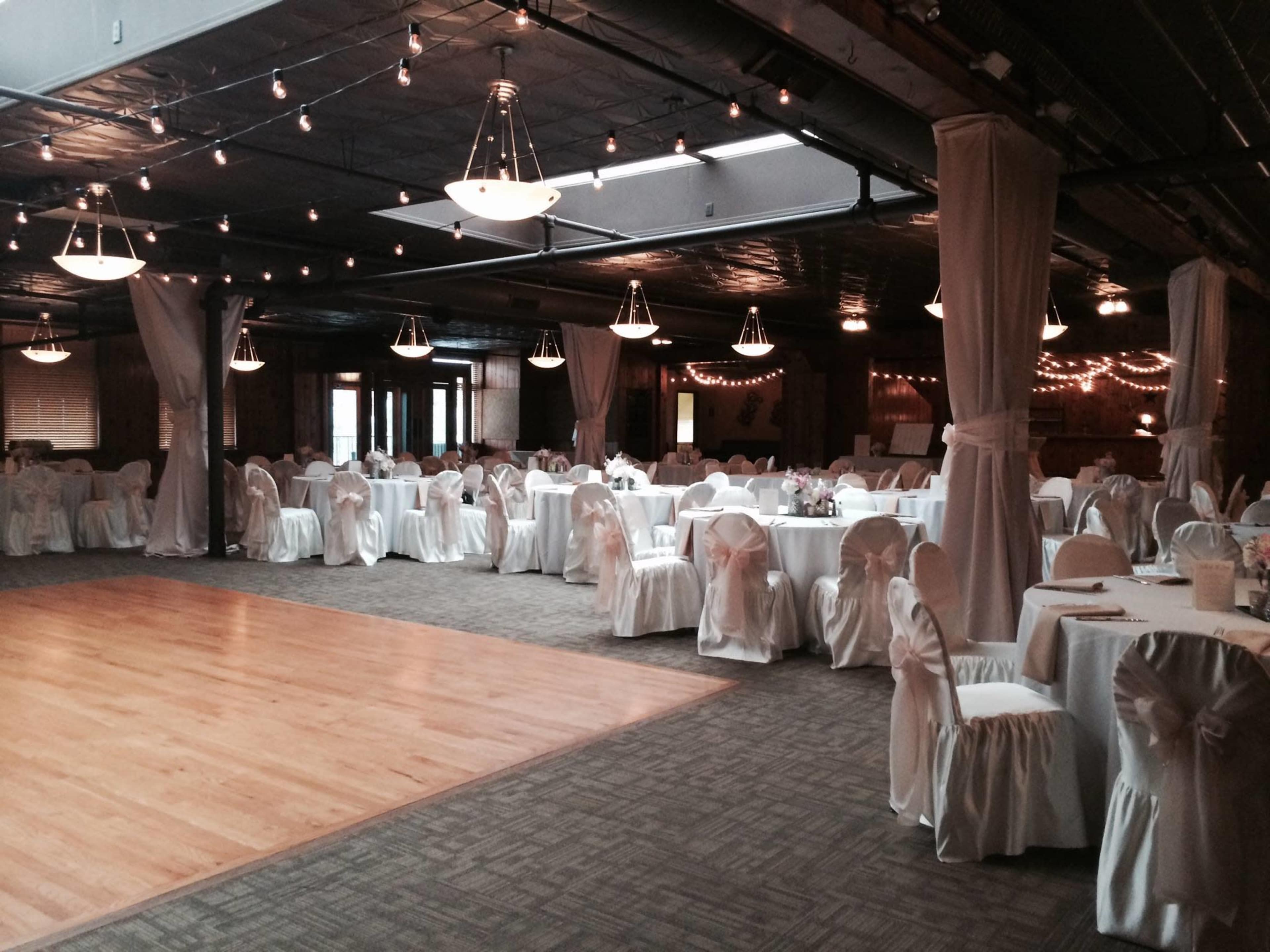 A banquet hall set up for a formal event, featuring round tables with white tablecloths and chairs draped in white fabric, illuminated by overhead lights and string lights.