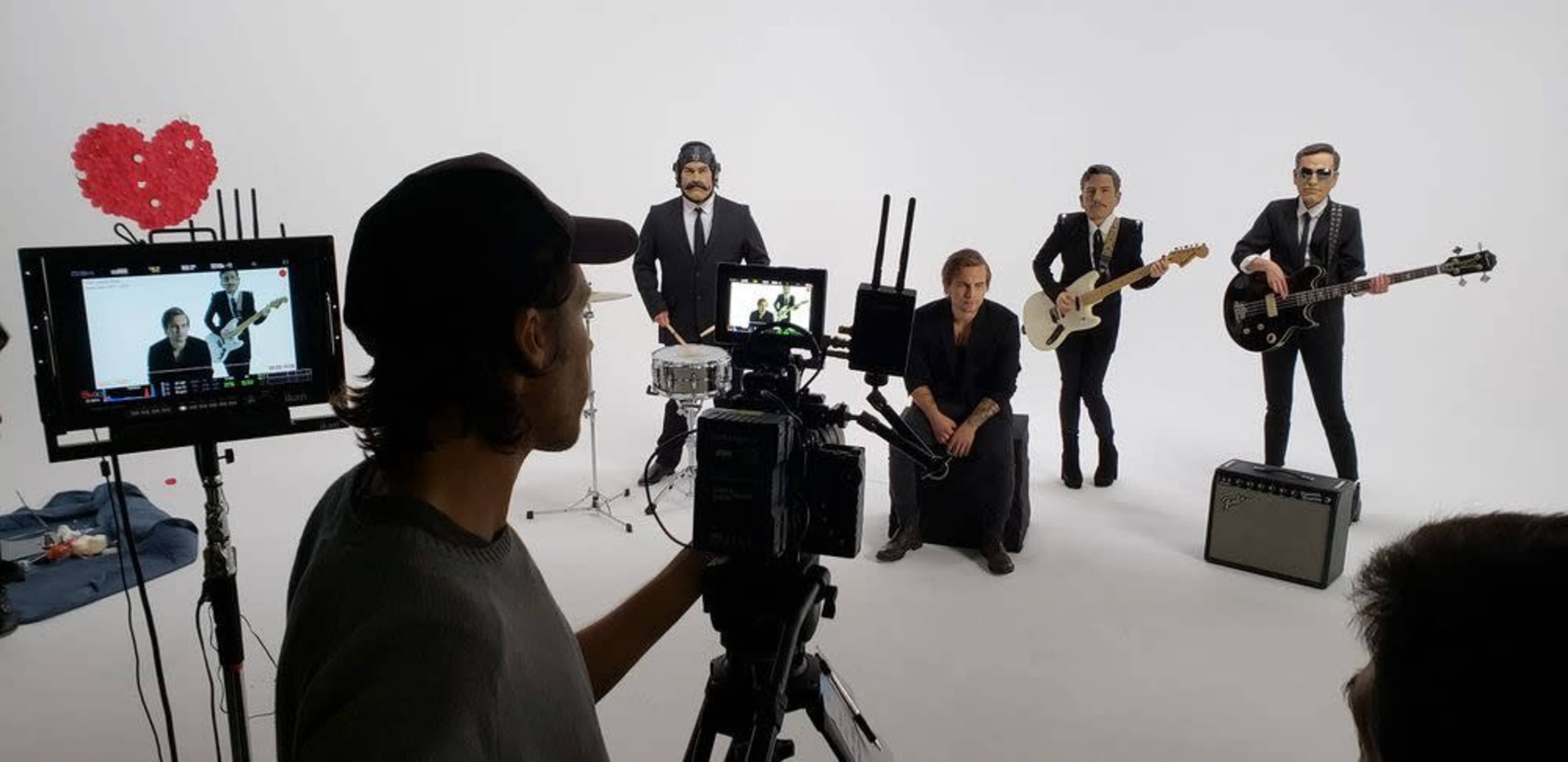 A film crew captures a band performing in a studio with a white backdrop and a heart-shaped decoration.