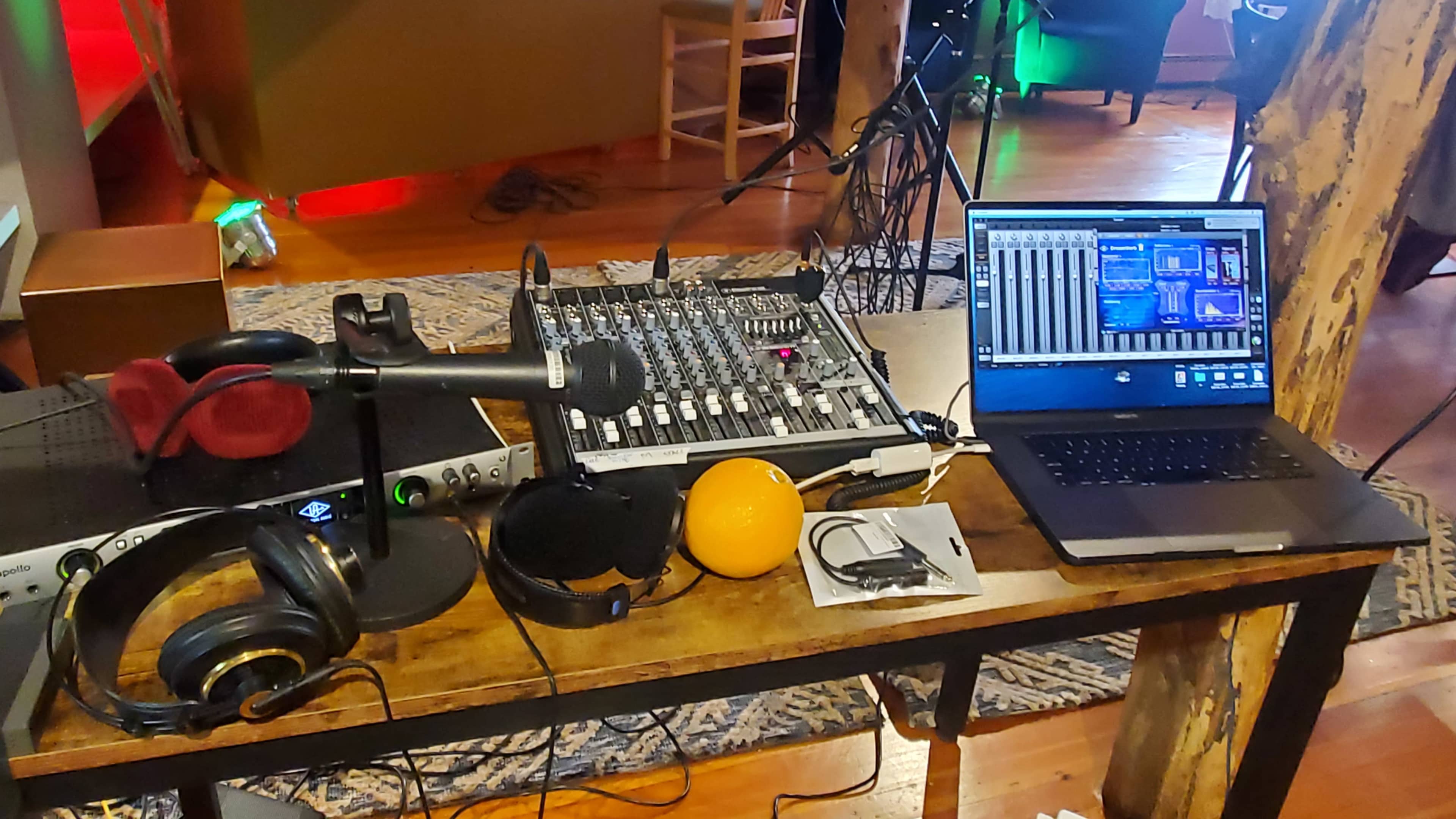 A mixing console, headphones, a microphone, and a laptop are displayed on a wooden table in a studio setting.
