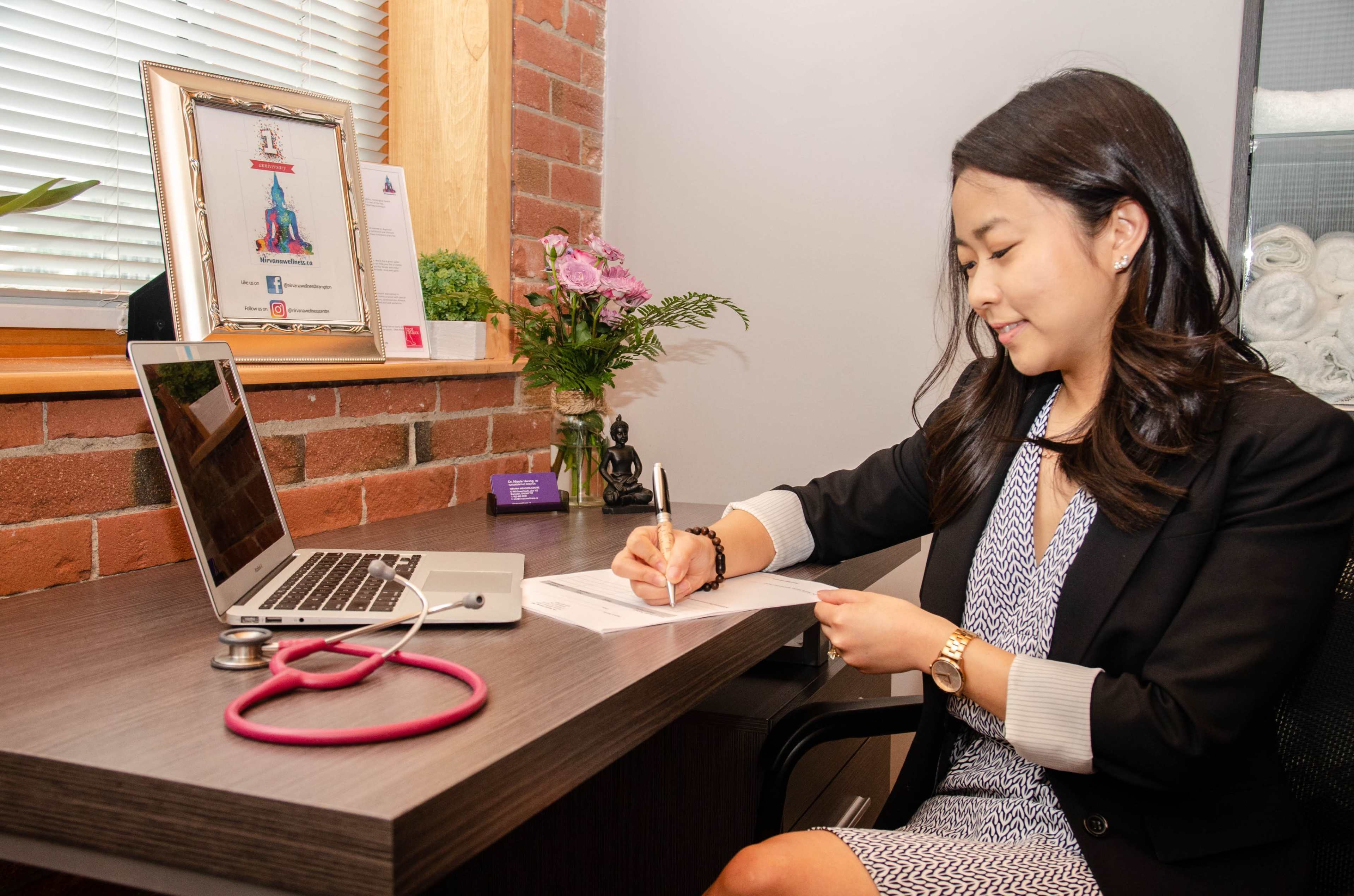A woman writes on a document at a desk with a laptop and a stethoscope nearby, surrounded by plants and framed certificates.